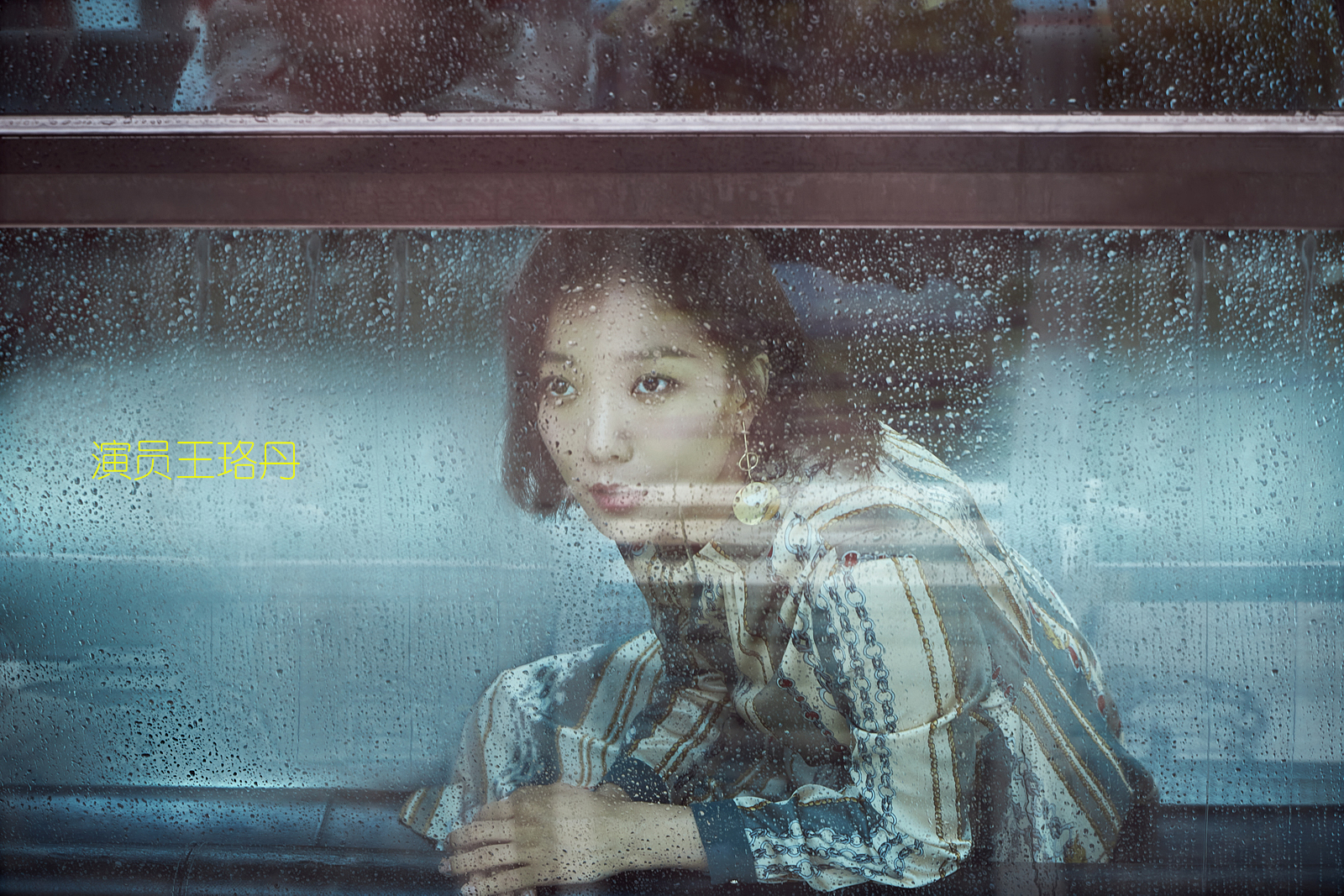 A woman with curly hair wearing a patterned blouse, sitting inside a vehicle, looking out through a rain-covered window. The window has droplets of water on it, and there is yellow text in Chinese next to her.