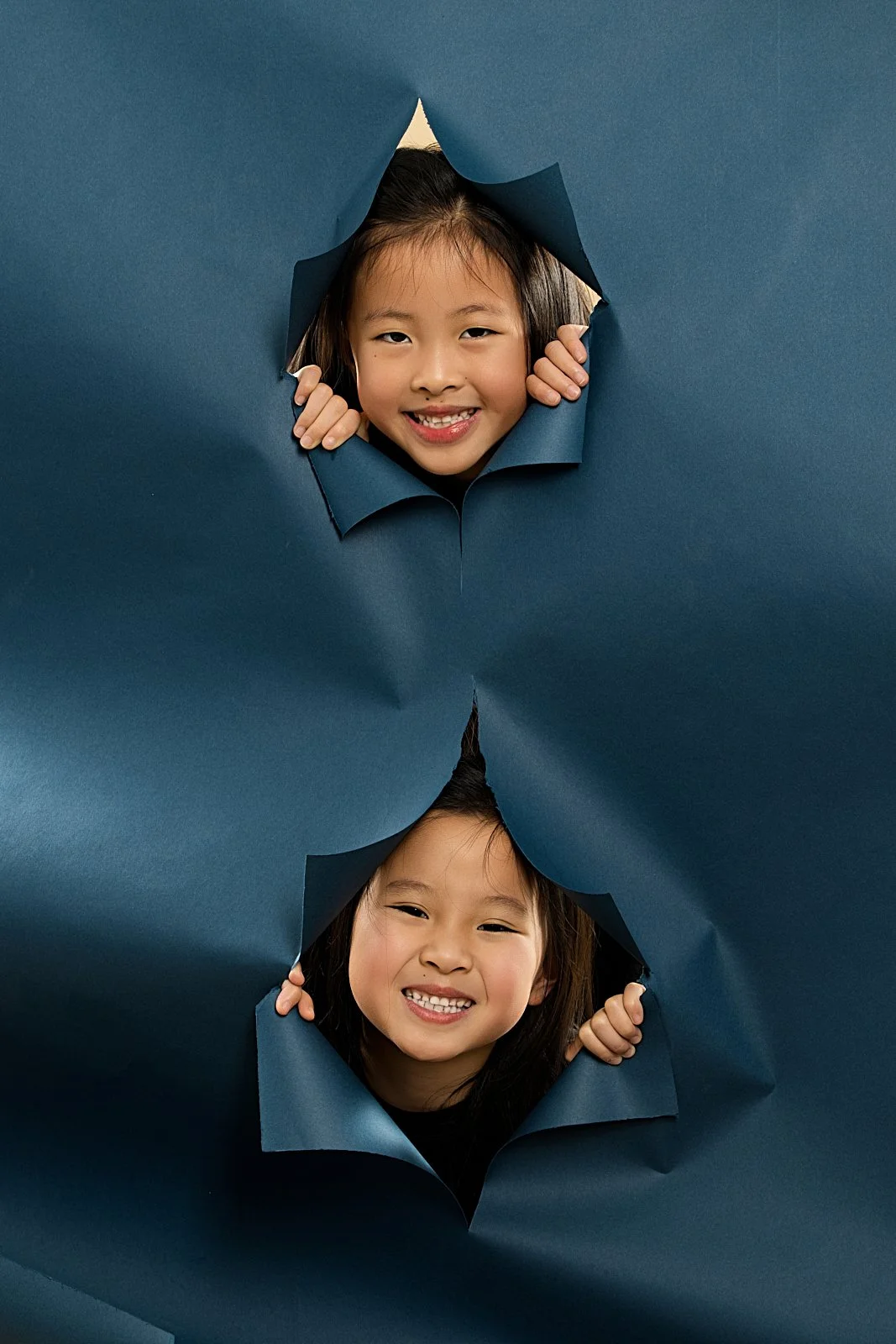 Two children peeking through torn blue paper with smiling faces.