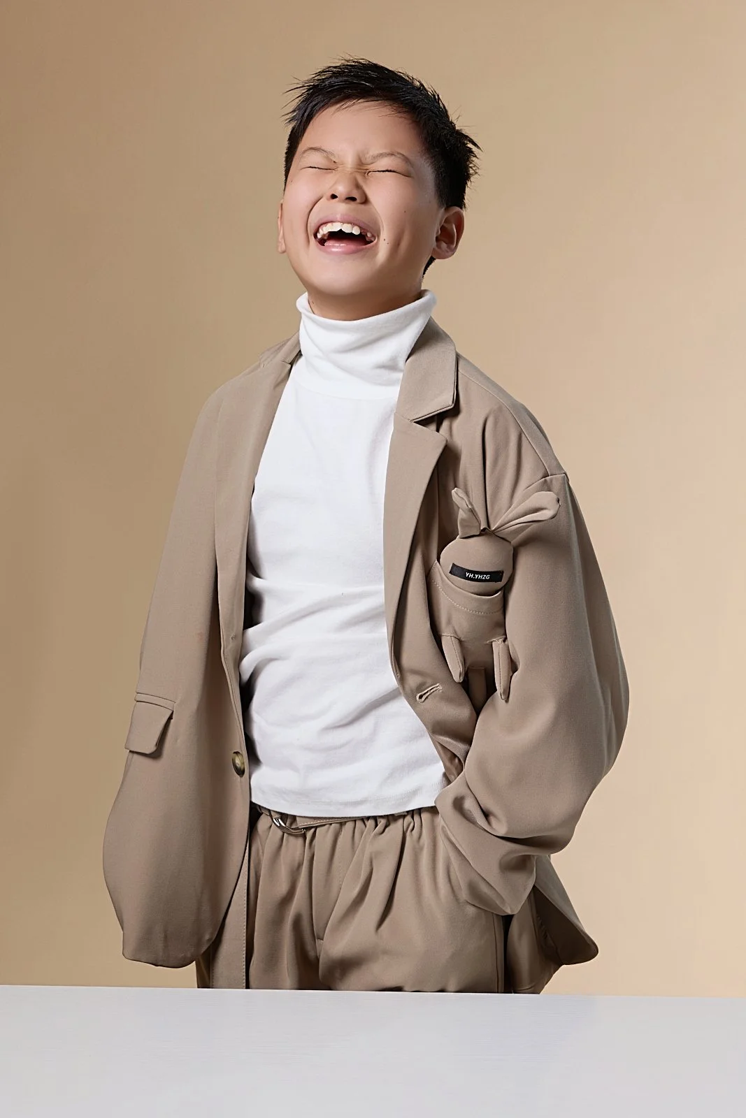 A young boy with short black hair, wearing a beige blazer over a white turtleneck and beige pants, standing against a plain beige background, with a joyful expression and eyes closed.