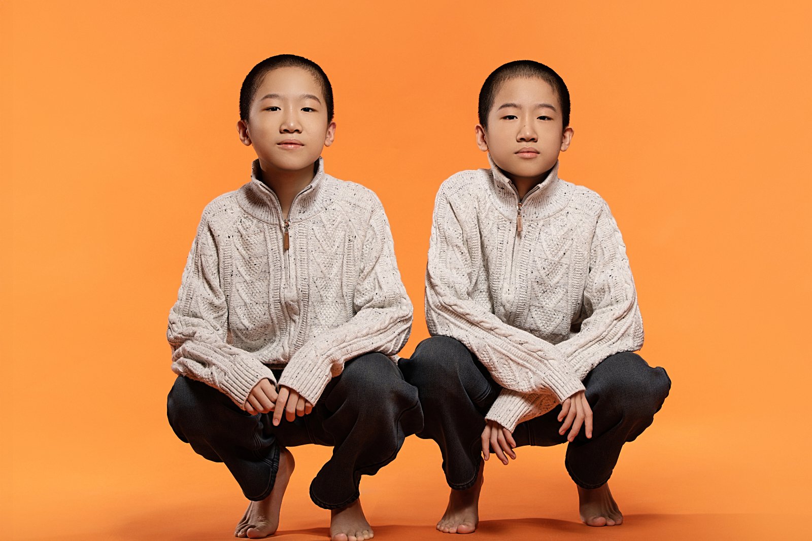 Two young boys sitting on the floor against an orange background. They are wearing matching cream-colored cable-knit sweaters and black pants, with serious expressions.