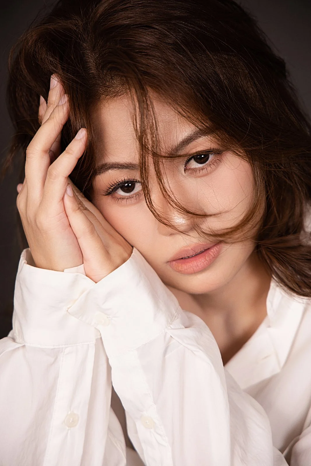 A woman with brown hair and natural makeup, dressed in a white shirt, resting her head on her hand and looking at the camera with a soft expression.