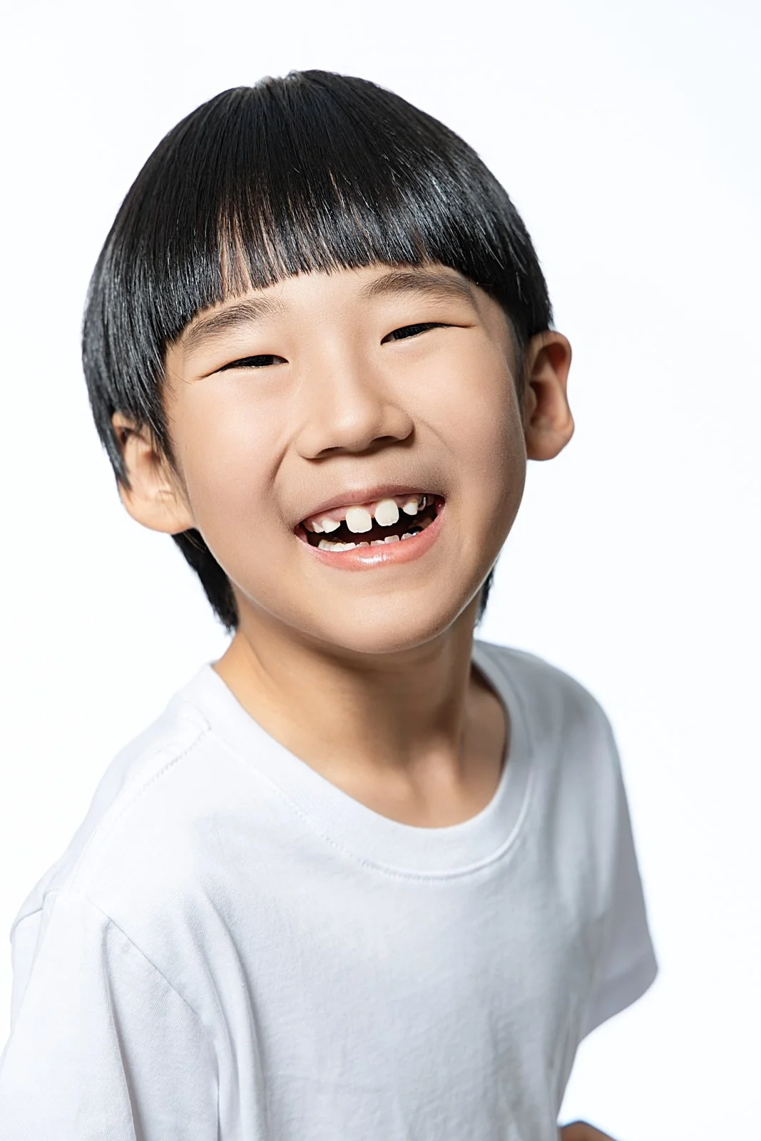 A young boy with black hair, missing front teeth, smiling and wearing a white shirt against a plain white background.