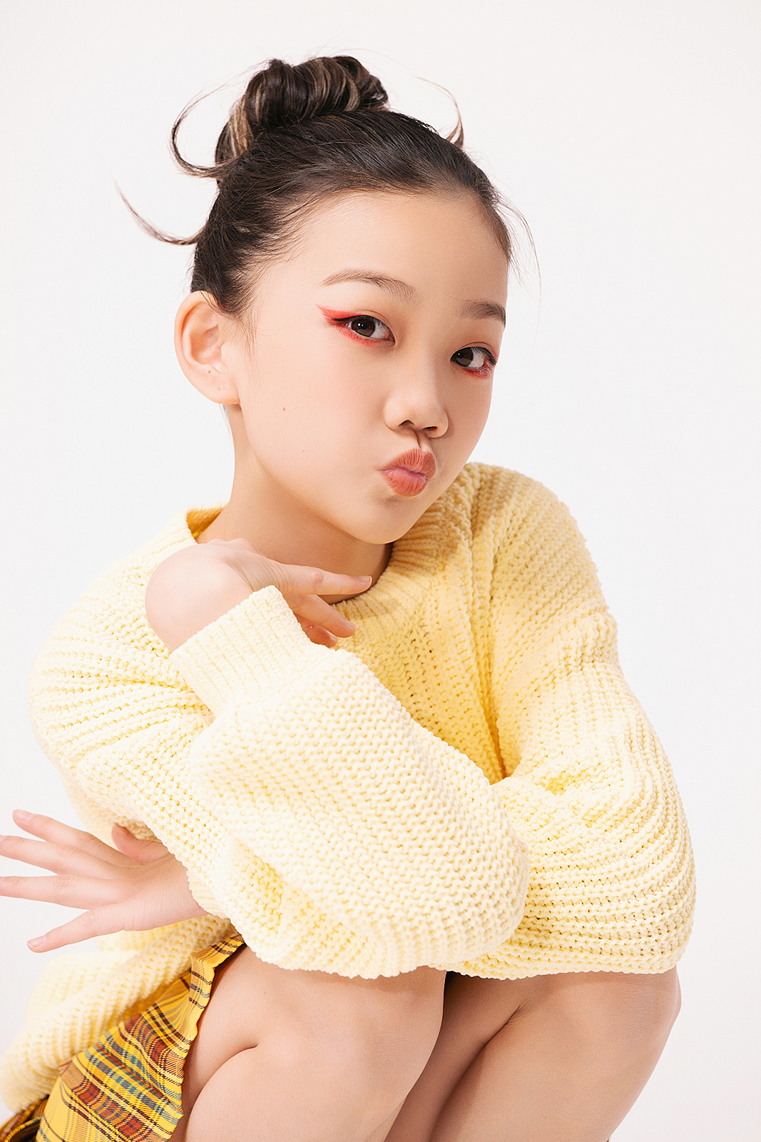 Young woman with dark hair tied up in a bun, wearing a yellow sweater and plaid skirt, squatting and making a playful face with puckered lips and red eyeliner.