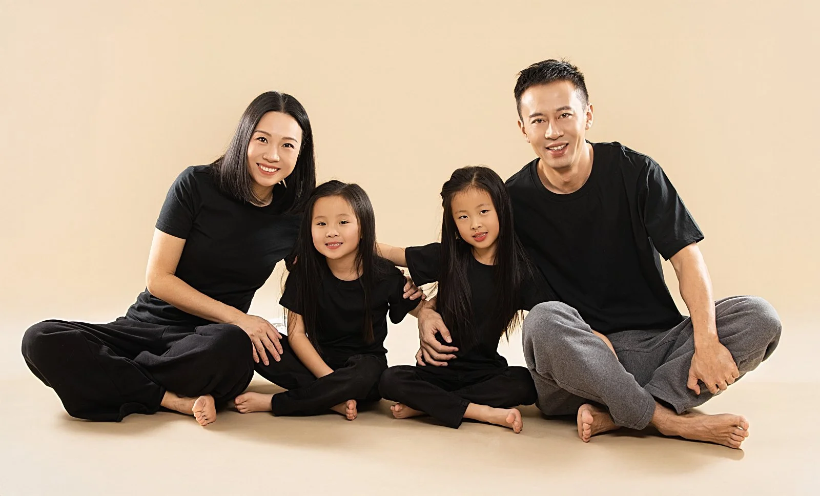 A family of four sitting on the floor against a beige background, all dressed in black and smiling.