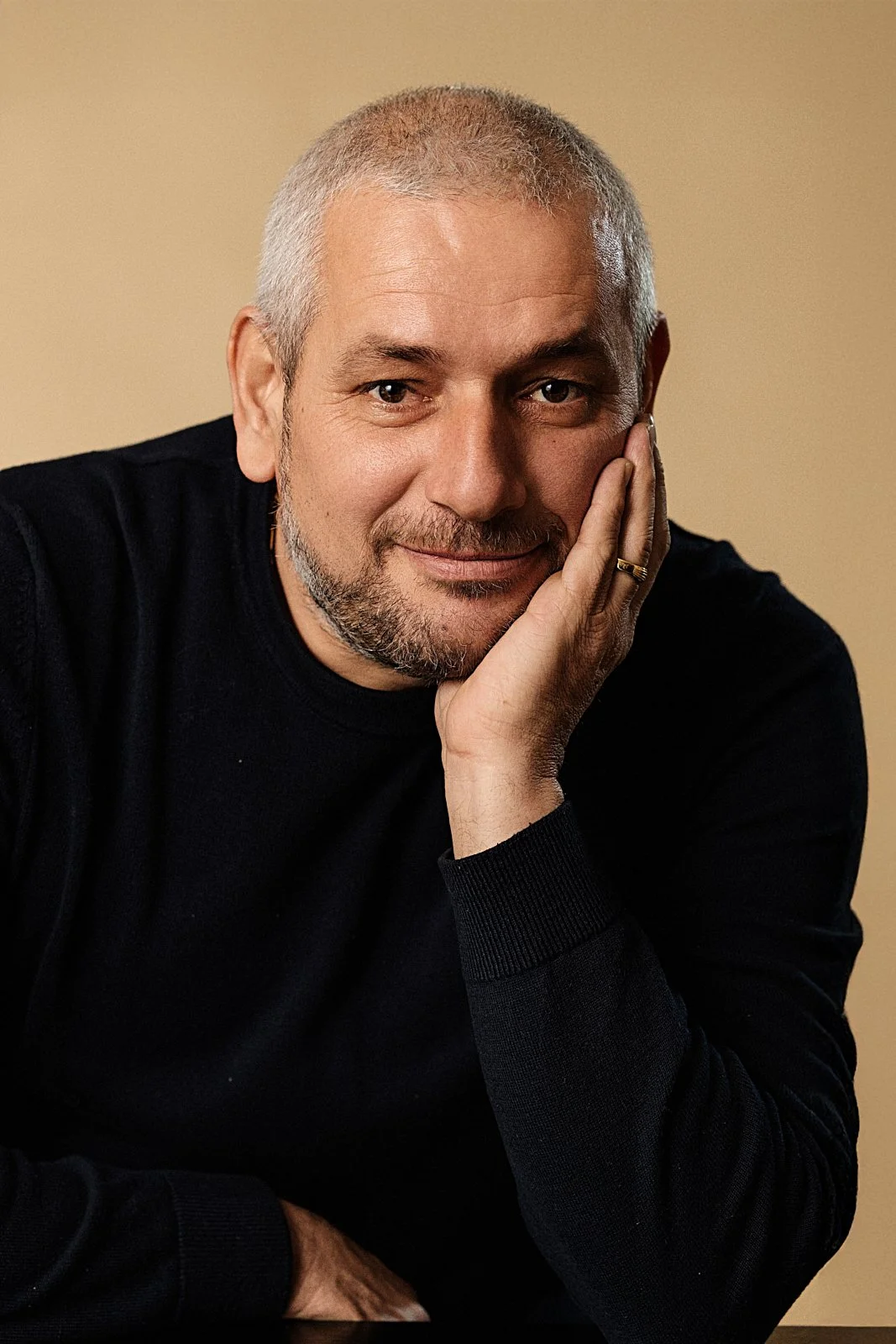Close-up of a middle-aged man with short gray hair and a beard, wearing a black sweater, resting his face on his hand, smiling softly against a beige background.