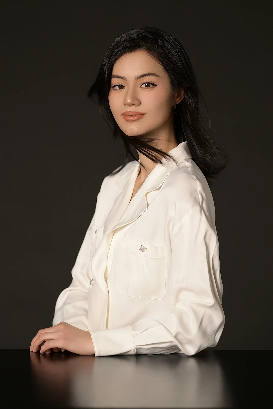 A woman with black hair wearing a cream silk blouse sitting at a dark table against a dark background.