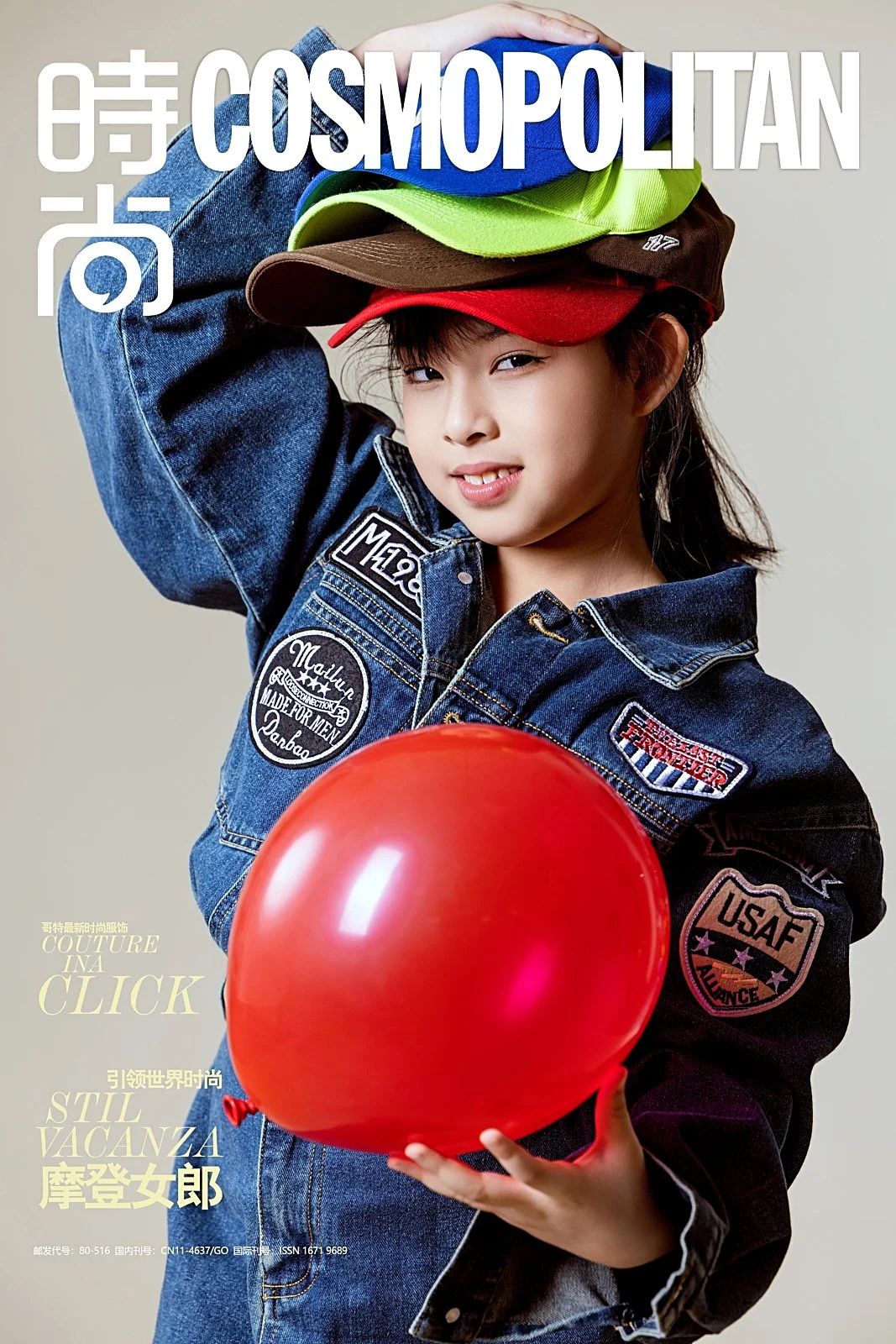 A young girl wearing a denim jacket and multicolored baseball caps stacking on her head, holding a red balloon, and posing for a fashion magazine cover.