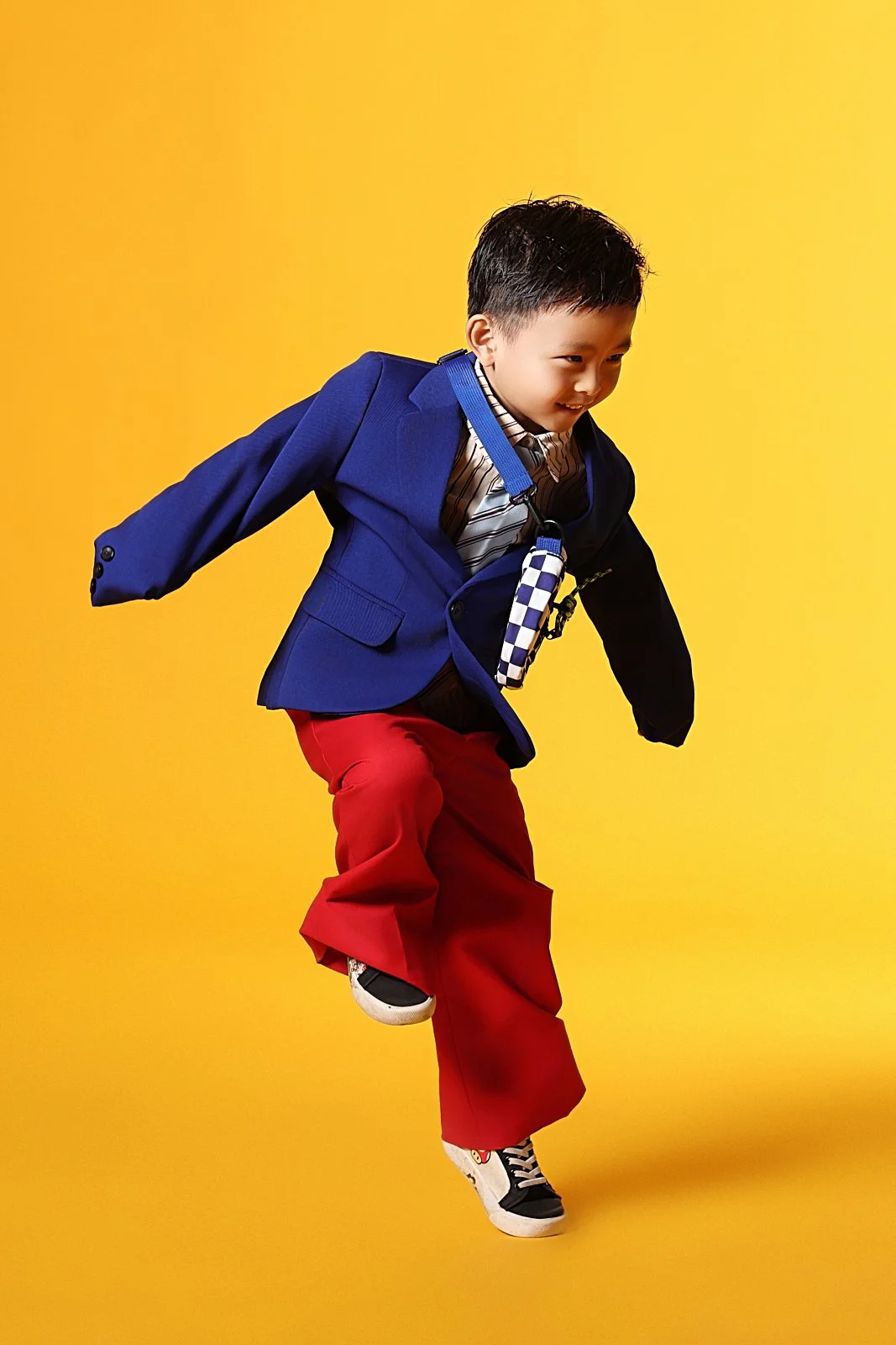 Young boy in a blue blazer, striped shirt, red pants, and sneakers, jumping with a bright yellow background.