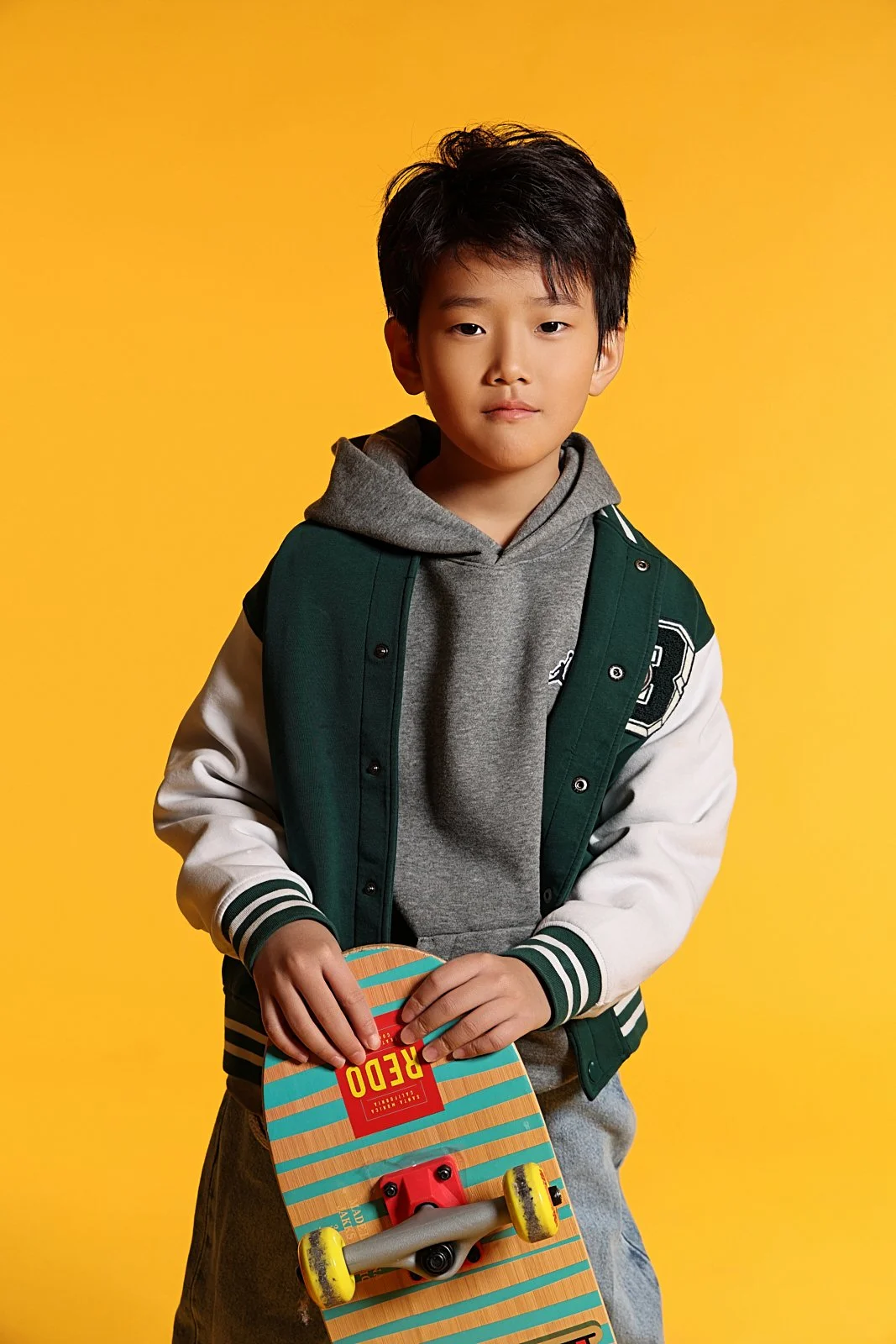 A young boy standing against a yellow background, holding a skateboard, wearing a gray hoodie and a green and white varsity jacket.