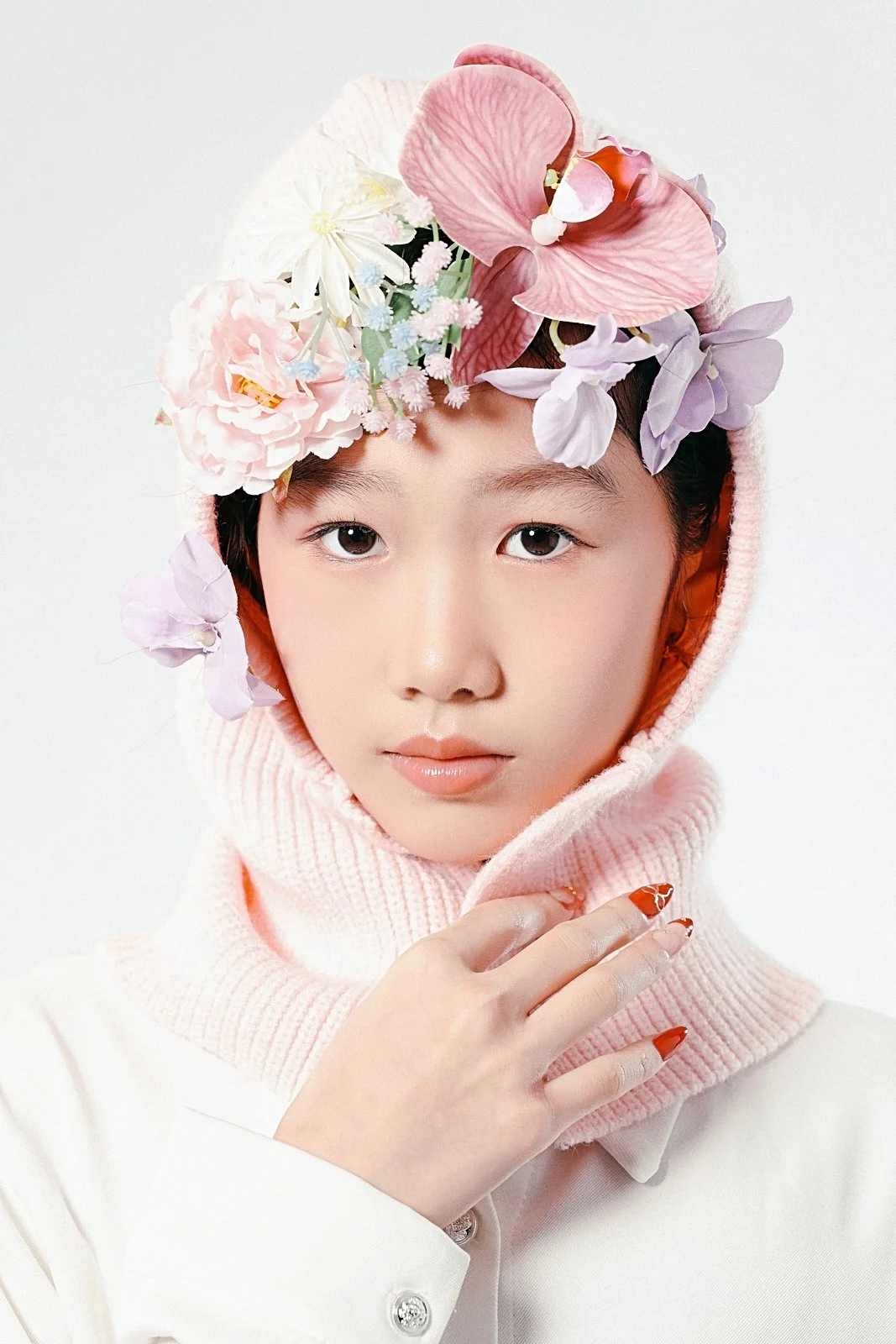A young person wearing a pink hoodie with a floral headpiece covering part of their head, featuring pink, purple, and white flowers. They have minimal makeup and are holding the collar of the hoodie with their right hand. The background is plain whit