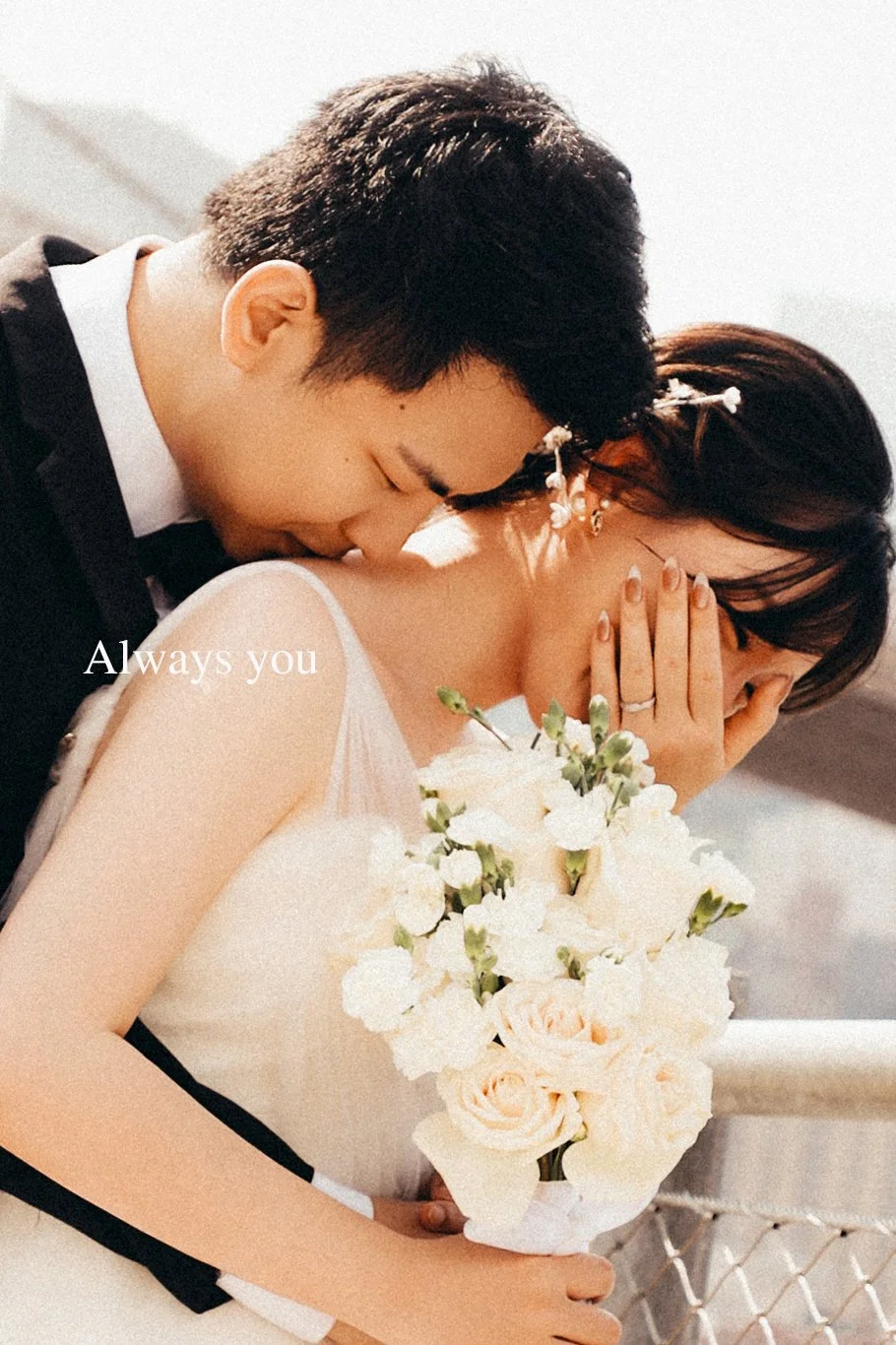 A couple dressed in wedding attire, with the groom kissing the bride on the shoulder. The bride holds a bouquet of white flowers, and the groom's hand rests on her shoulder. The bride is covering her face with her hand, and the word "Always you" is o