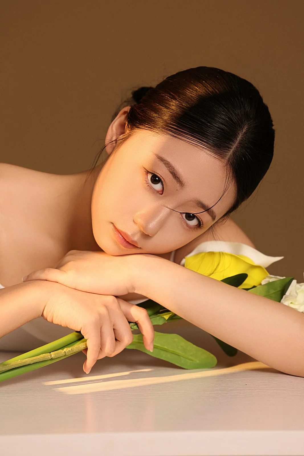 A woman with dark hair lying on a table, resting her head on her arm, surrounded by yellow and white flowers.