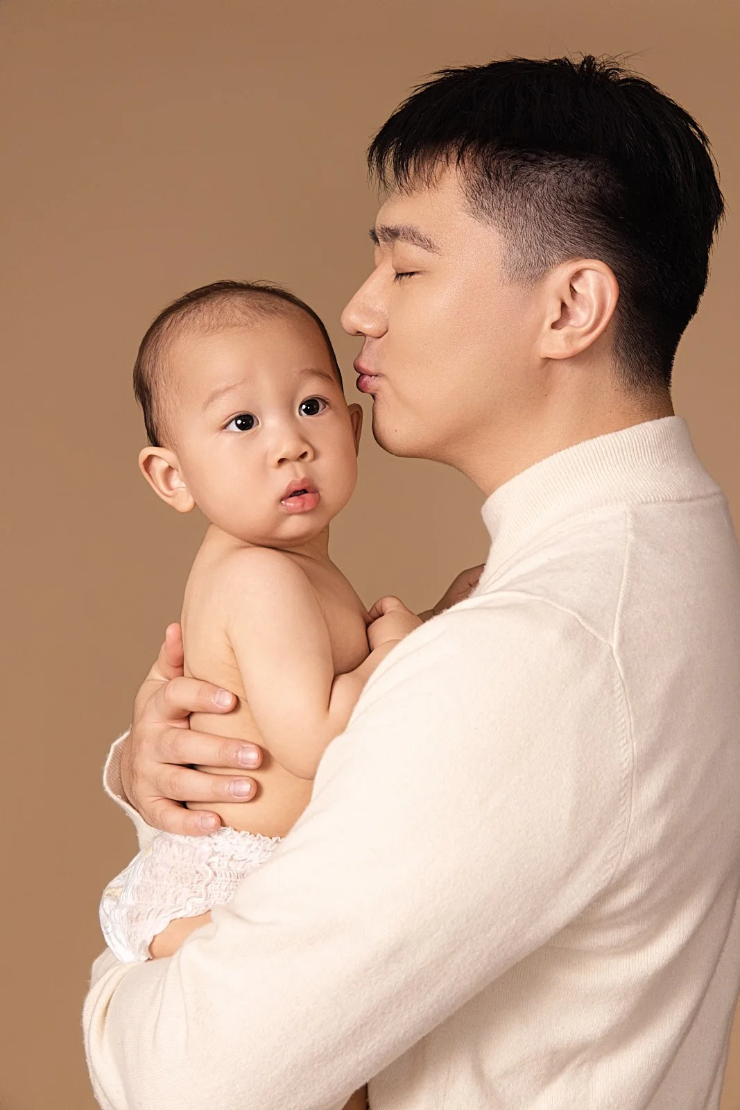 A man holding a young baby, giving a gentle kiss on the forehead. The baby looks surprised and is wearing only a diaper. The background is plain beige.