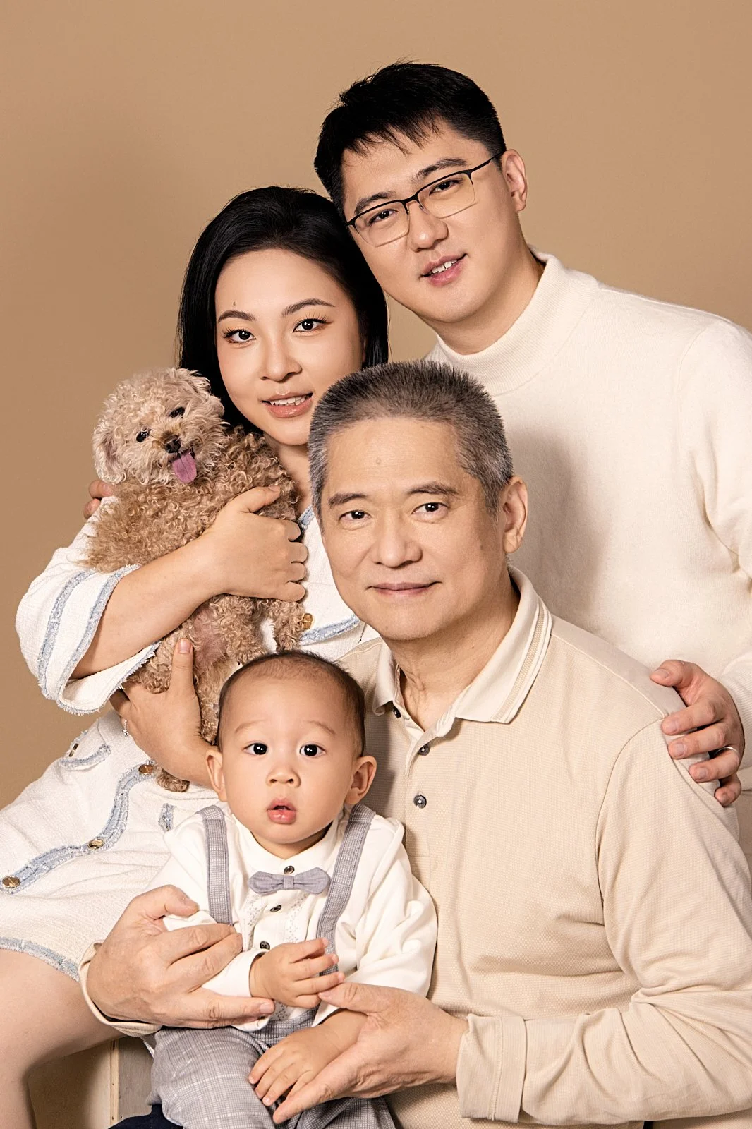 A multi-generational Asian family portrait with grandparents, parents, a young child, and a woman holding a small dog against a neutral background.
