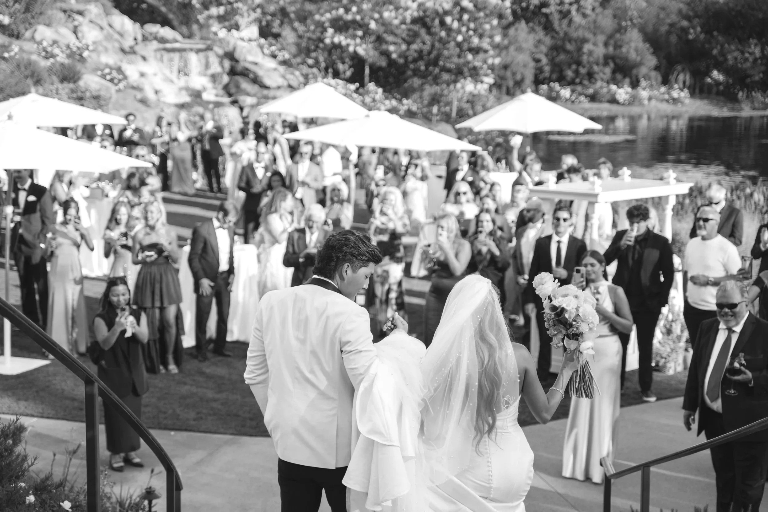 Black and white photo of a wedding reception outdoors by a lak. The bride with long hair in a white dress and veil, holding a bouquet, is walking with the groom in a white suit. Guests surround them, taking photos and watching, under umbrellas near the water