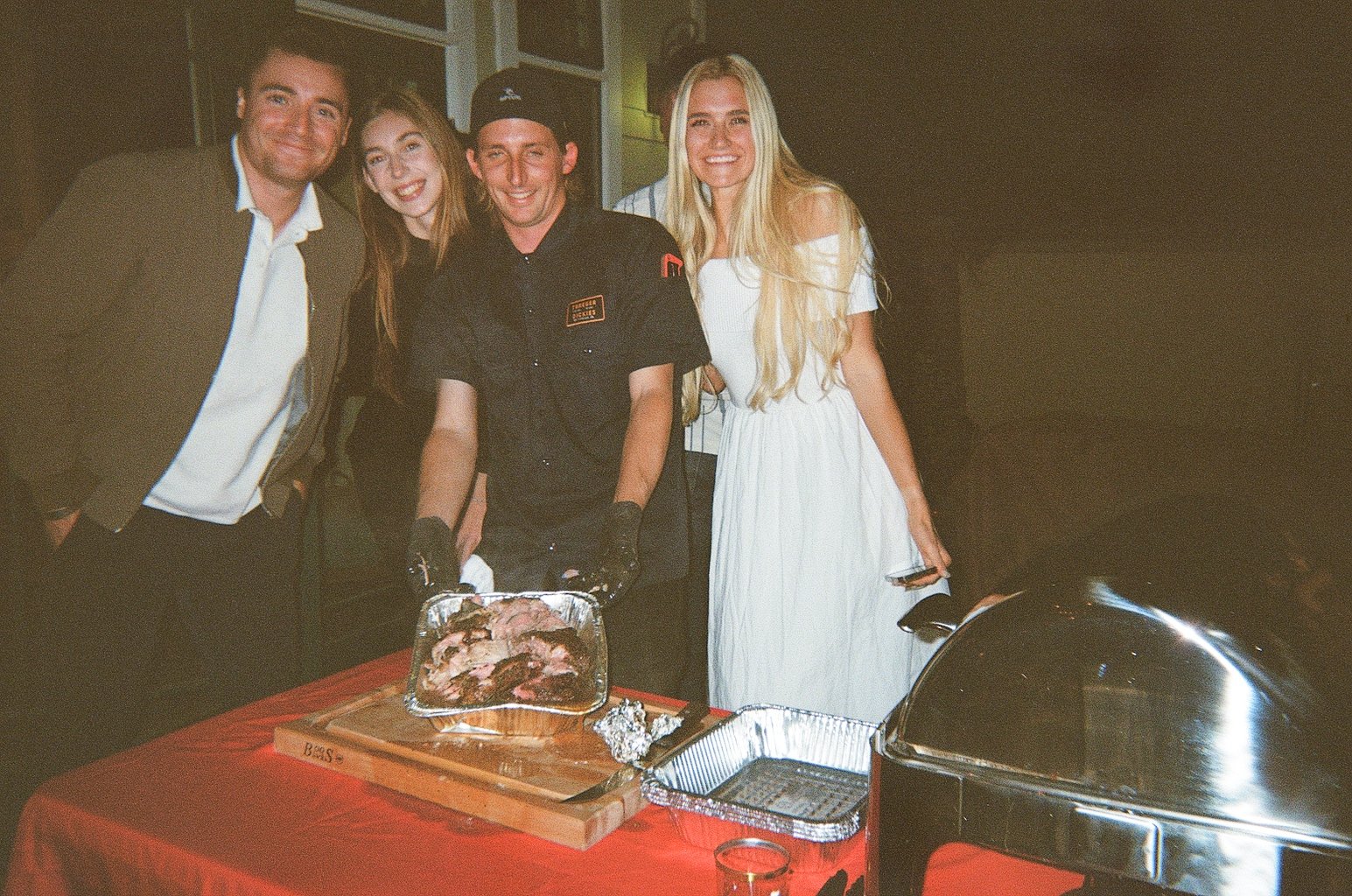 Bride and groom standing behind a table with cooked meat in aluminum trays at a social gathering.
