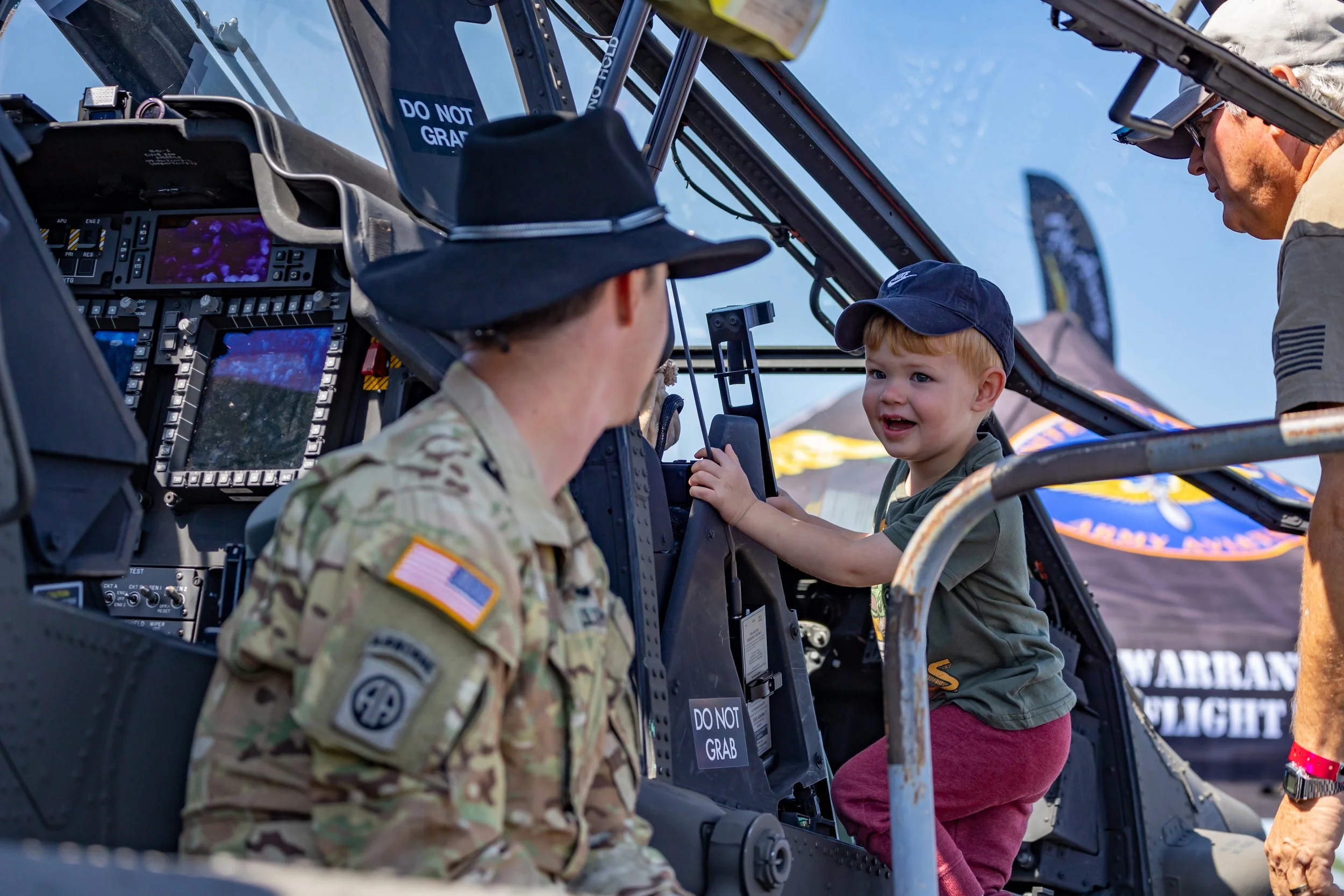 A young smiling boy sitting inside the cockpit of a military helicopter, talking to a woman in camouflage military uniform and other person standing outside. The boy is wearing a navy cap, and the military personnel is wearing a black hat.