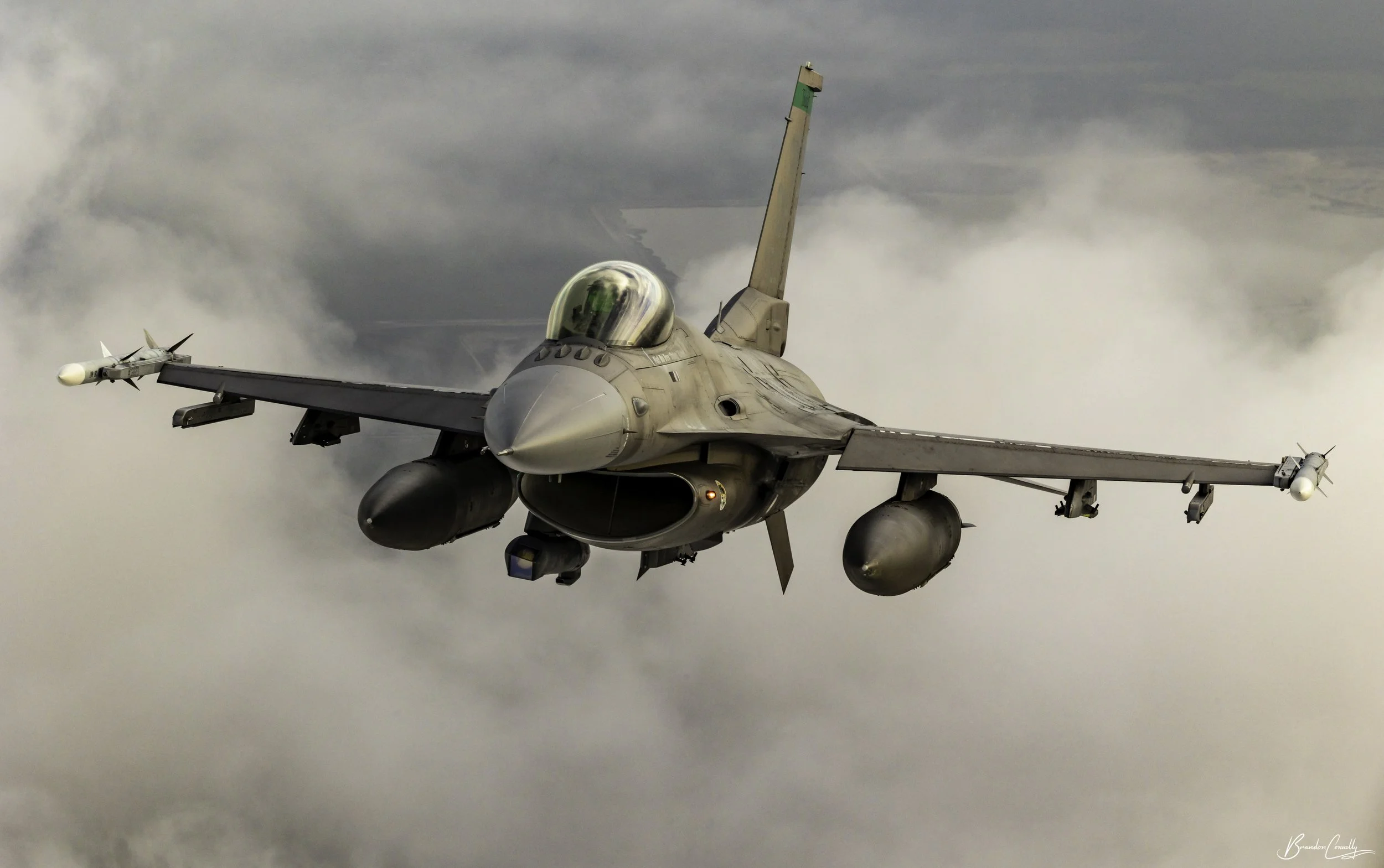 An F-16 fighter jet flying through cloudy skies, equipped with external fuel tanks and missiles under its wings.
