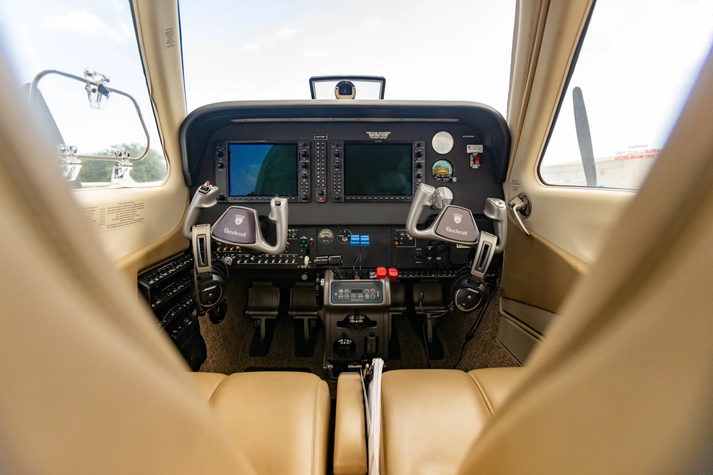Aircraft cockpit with dual control yokes, instrument panels, and digital screens, beige interior, and front windows showing the sky.