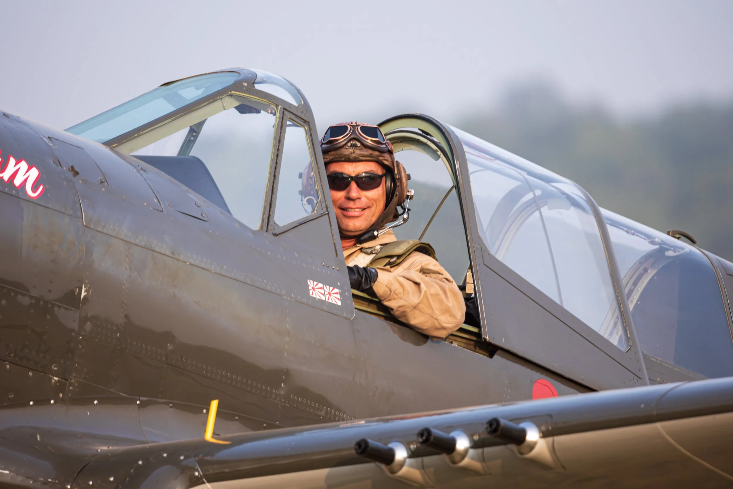 A pilot sitting in the cockpit of a vintage military aircraft, wearing sunglasses, a flight helmet, and a tan flight jacket, smiling at the camera.