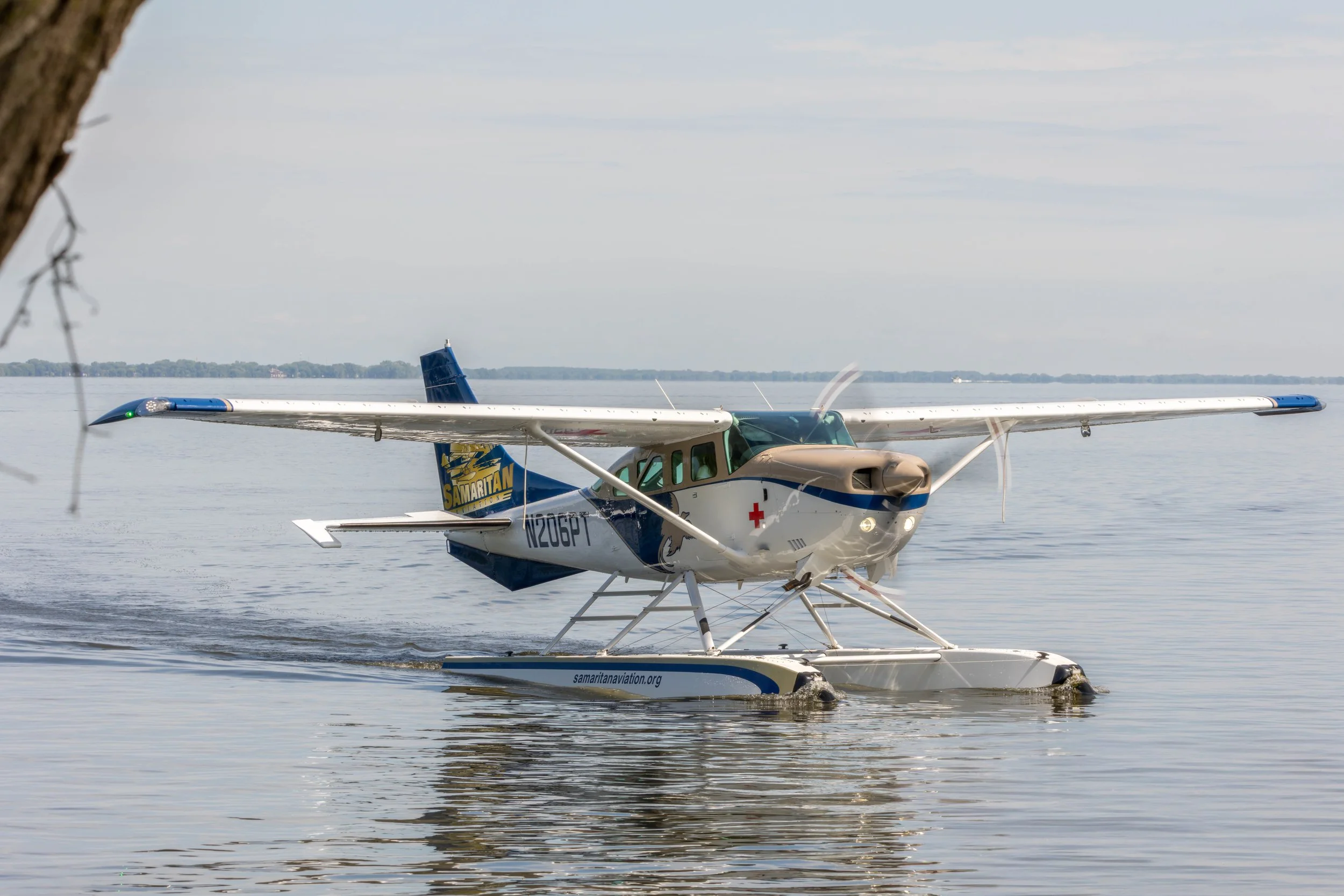 A seaplane flying low over calm water, with a tree branch partially visible on the left side, and distant shoreline with trees in the background.