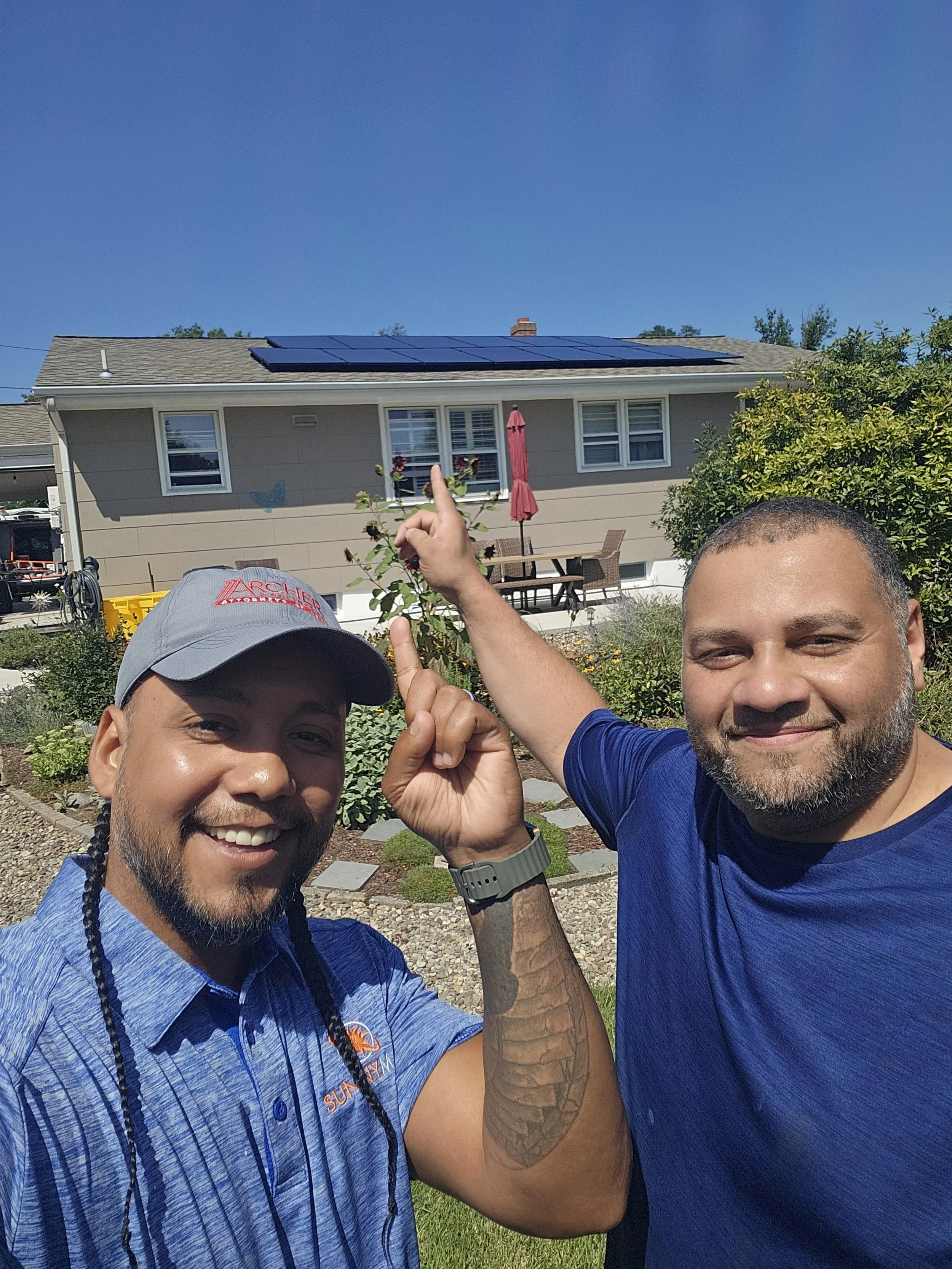 Jose the Sun Hunter and a homeowner smile and point proudly to new rooftop solar panels installed on a single-story NJ home. Clear skies and solar success highlight clean energy and customer satisfaction.