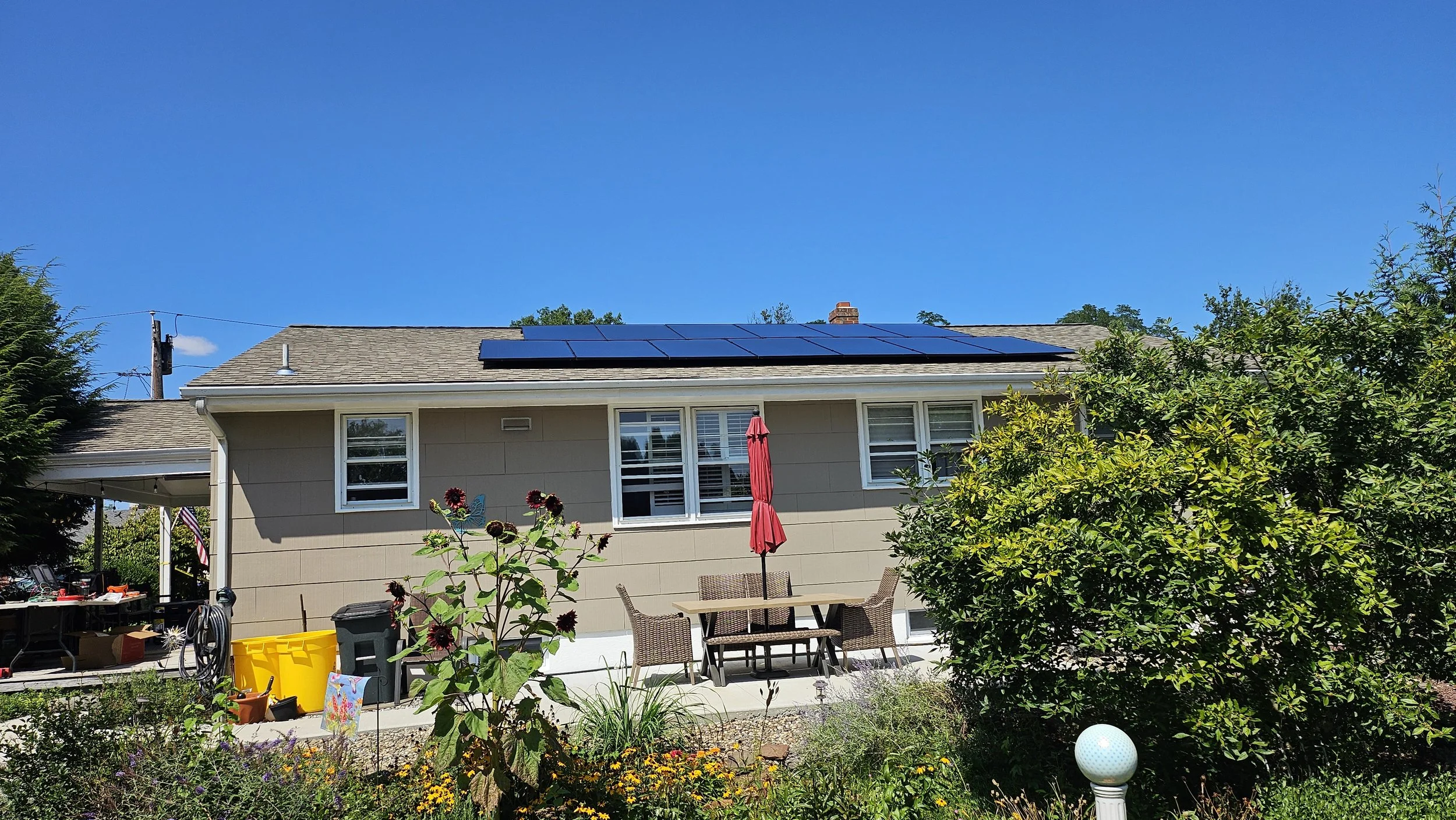 Backyard view of a single-story NJ home featuring a full rooftop solar panel system under a clear blue sky. Patio furniture, garden flowers, and clean installation highlight residential solar energy benefits.