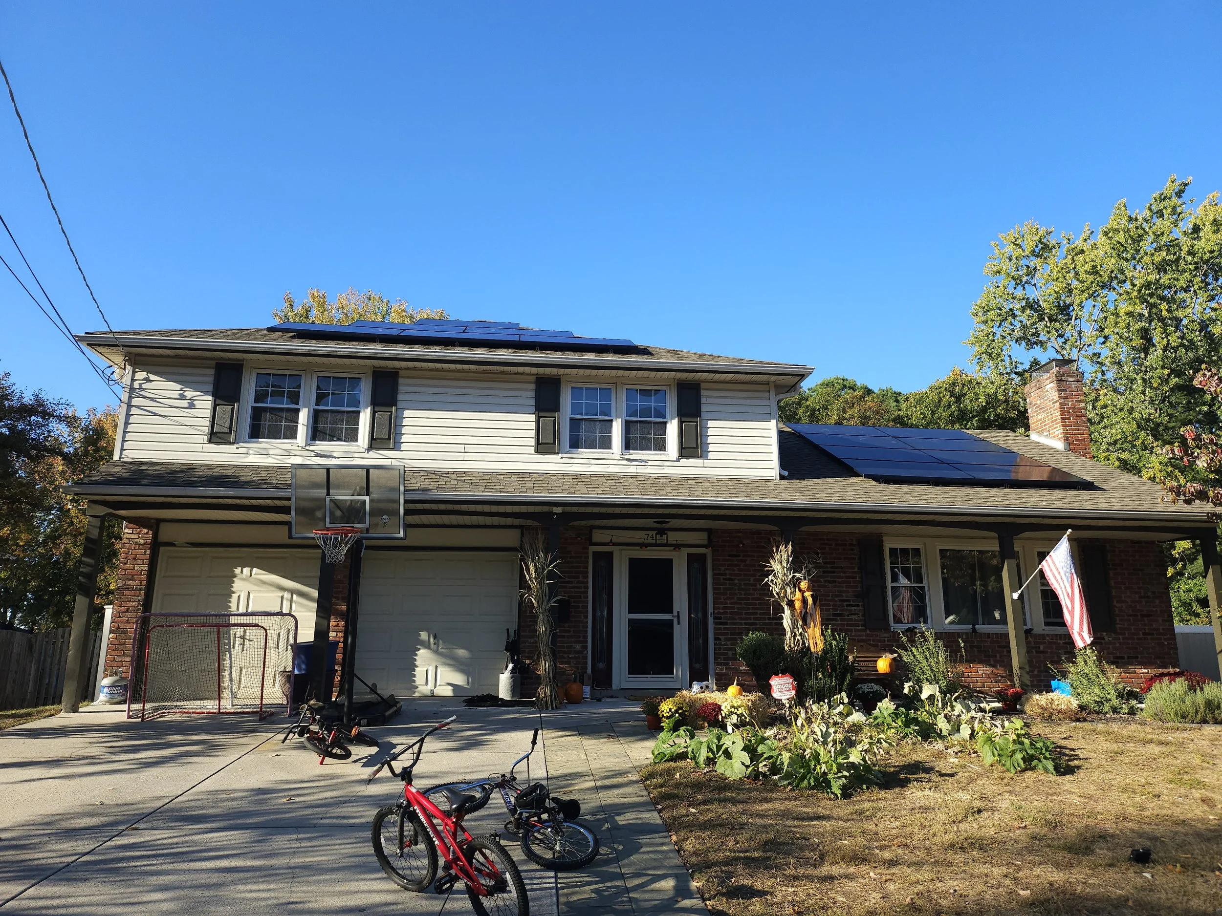 Front view of a two-story NJ home with a full rooftop solar panel installation under a clear blue sky. American flag, kids' bikes, and fall decorations highlight a family-friendly solar-powered household.