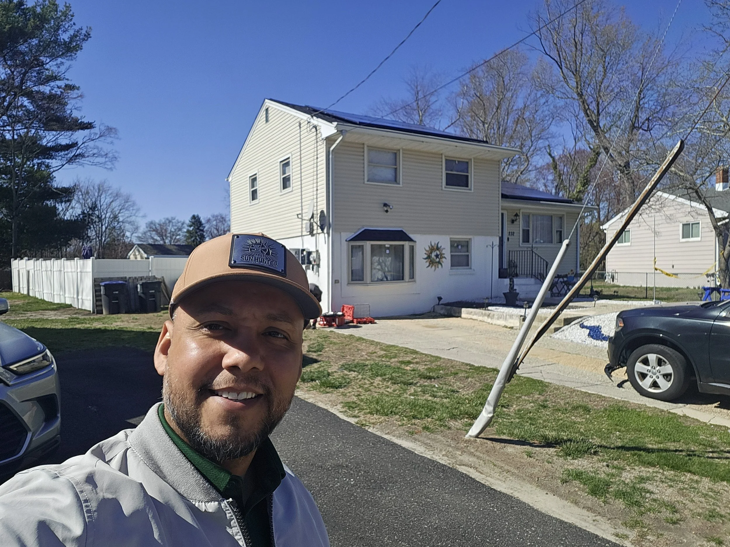 Smiling solar consultant wearing a Sun Hunter cap stands in front of a two-story beige house with newly installed rooftop solar panels on a sunny day.