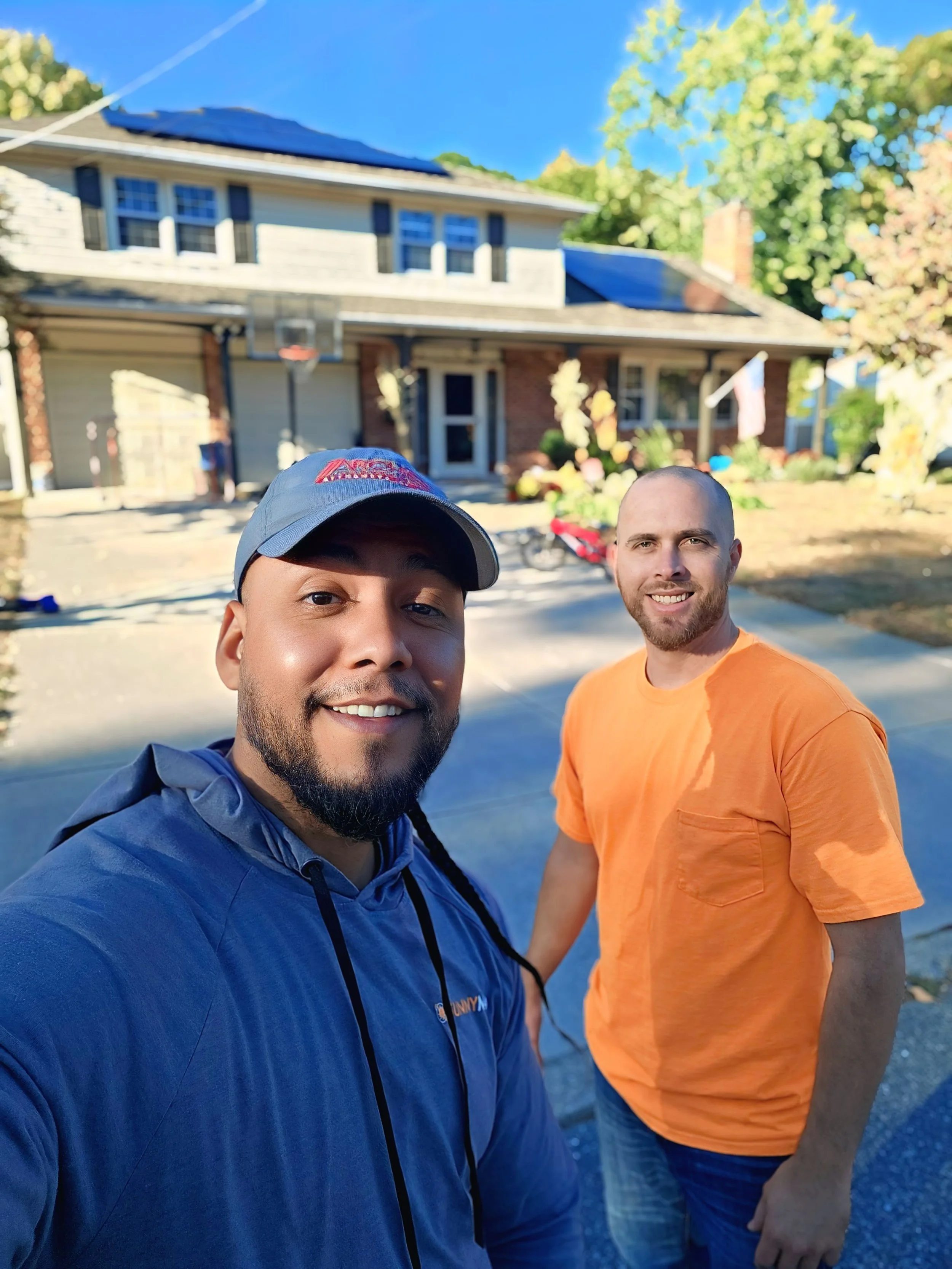 Jose the Sun Hunter smiling with a satisfied homeowner in front of a two-story NJ home with new rooftop solar panels. Clear skies and solar installation highlight clean energy success and local savings.