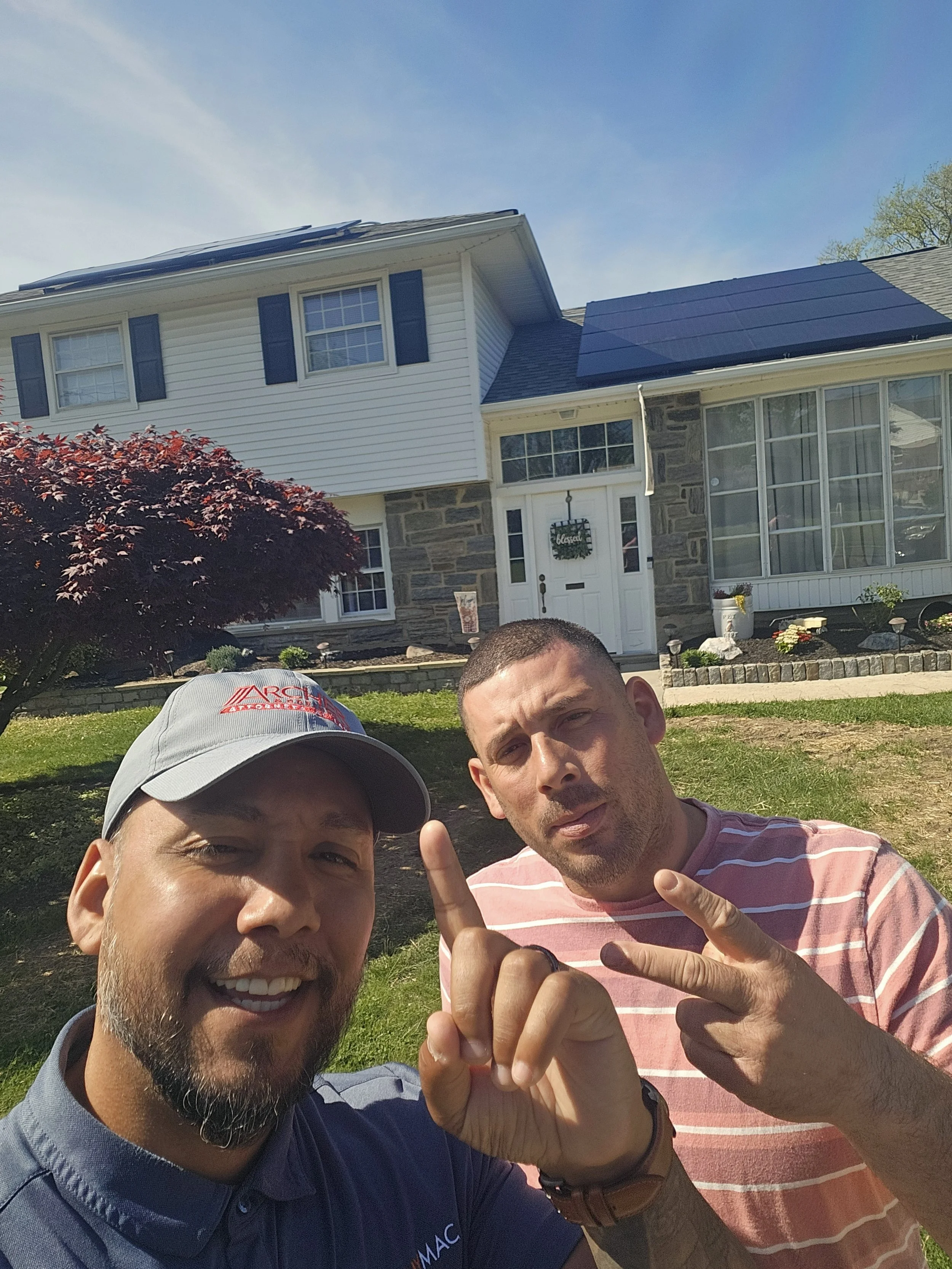 Jose the Sun Hunter and a proud homeowner pose in front of a two-story NJ home with new solar panels installed on both roof sections. Celebrating solar savings and energy independence with clear skies and high spirits.