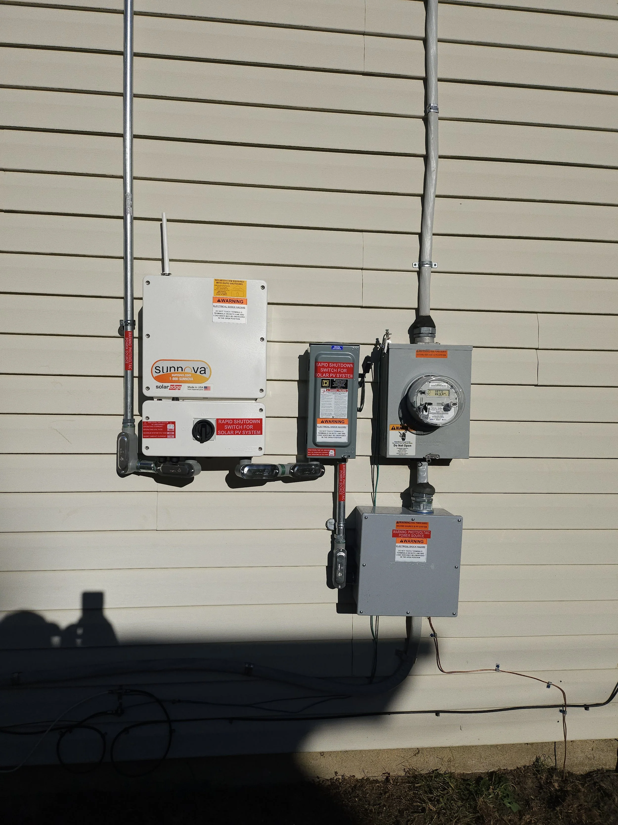 Close-up of a Sunnova solar inverter system, utility meter, and electrical components mounted on the exterior wall of a solar-powered home in New Jersey.