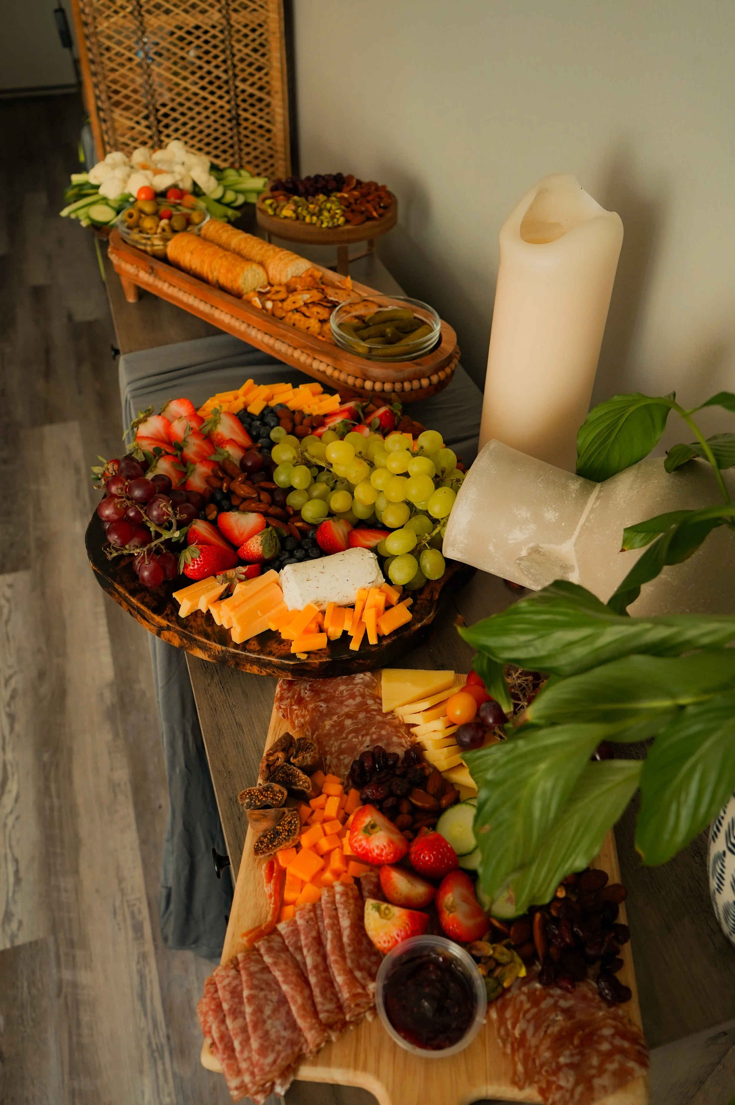 A long table filled with a variety of snacks, cheeses, fruits, nuts, cured meats, and a lit white candle. The table is decorated for a gathering or party.