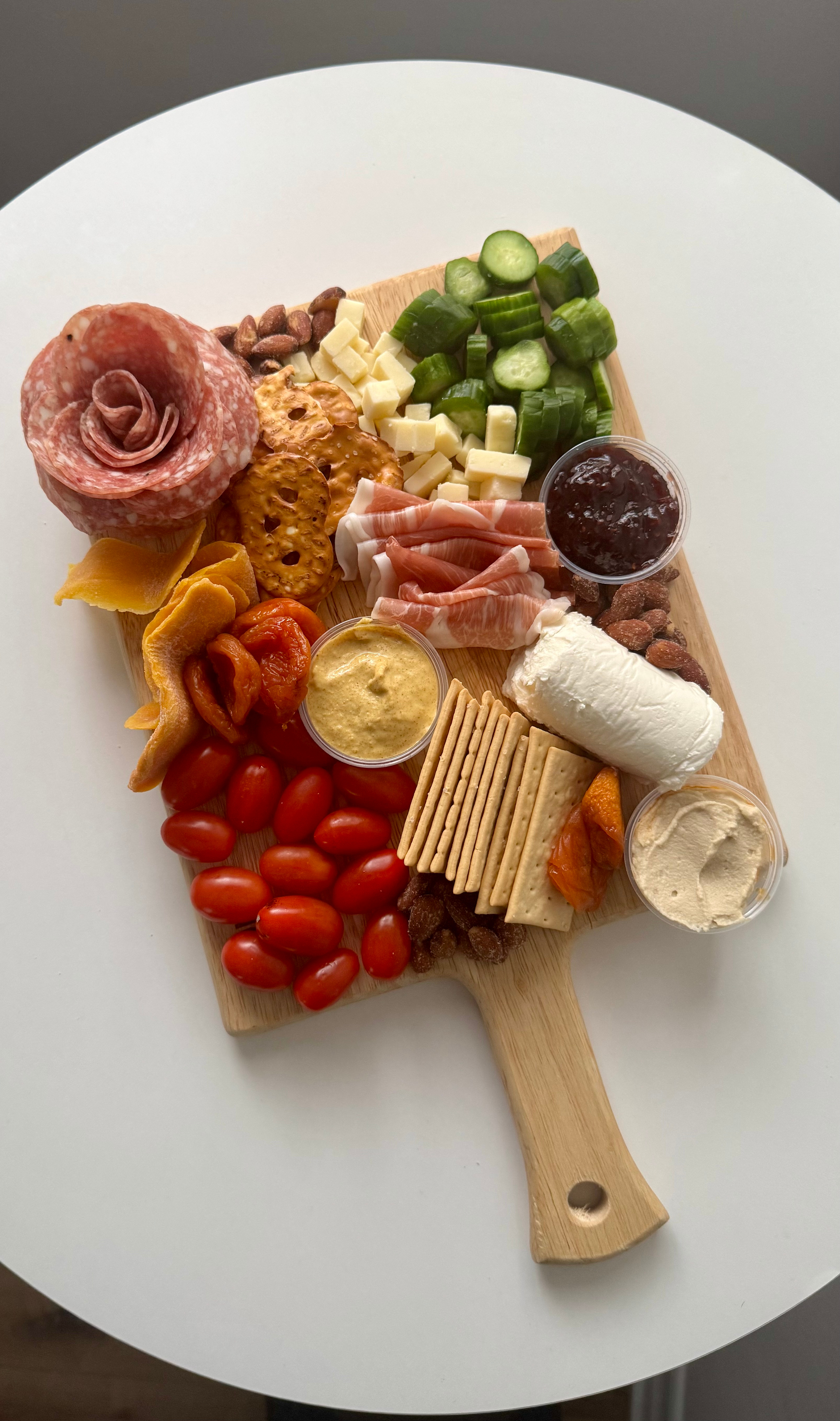 Charcuterie board with cherry tomatoes, cheese, crackers, sliced cucumber, salami, prosciutto, almonds, pretzels, jam, mustard, and a spread.