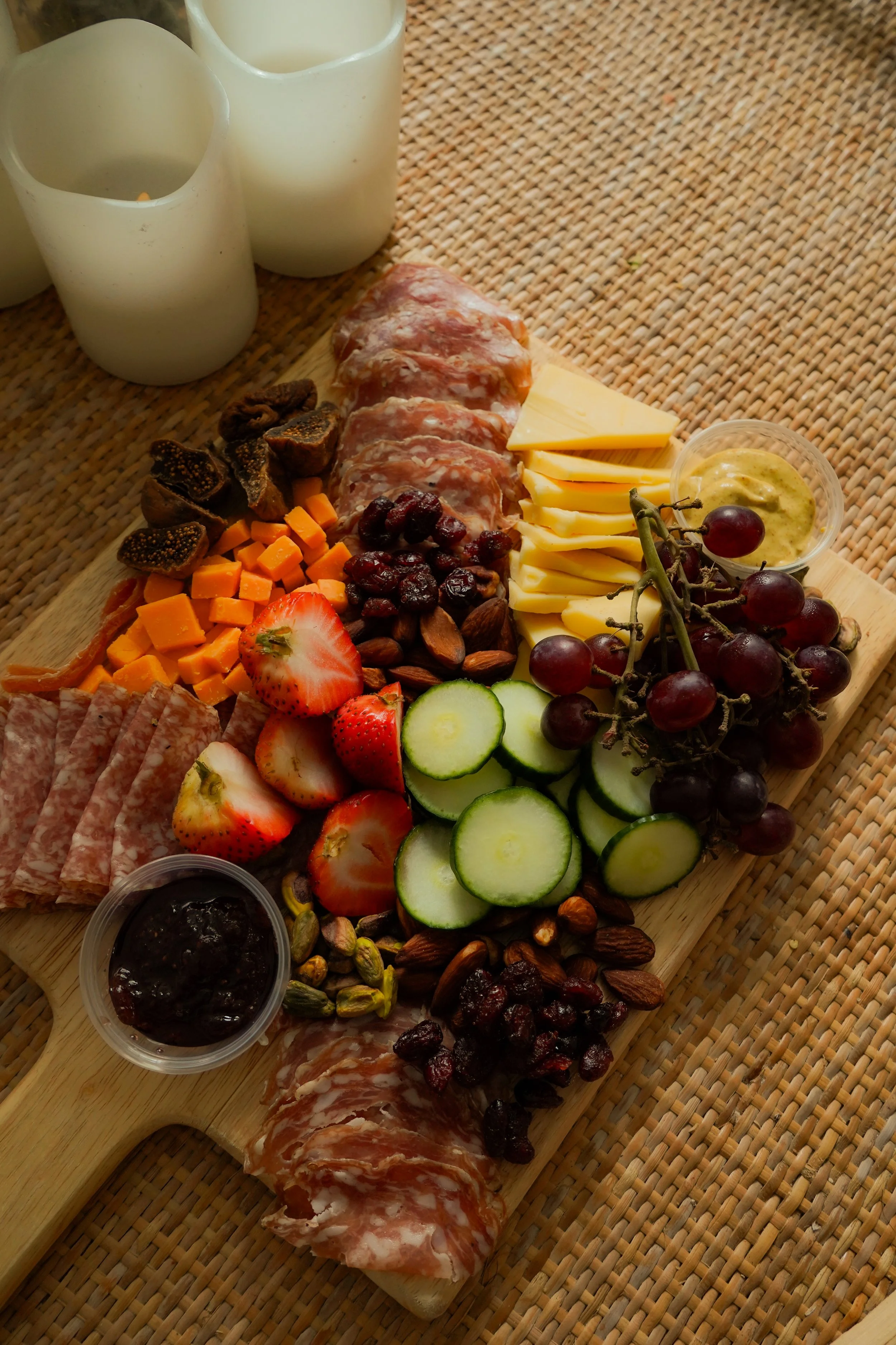 A charcuterie board with cheese, cured meats, grapes, strawberries, figs, dried fruits, nuts, and a container of jam, on a woven placemat.