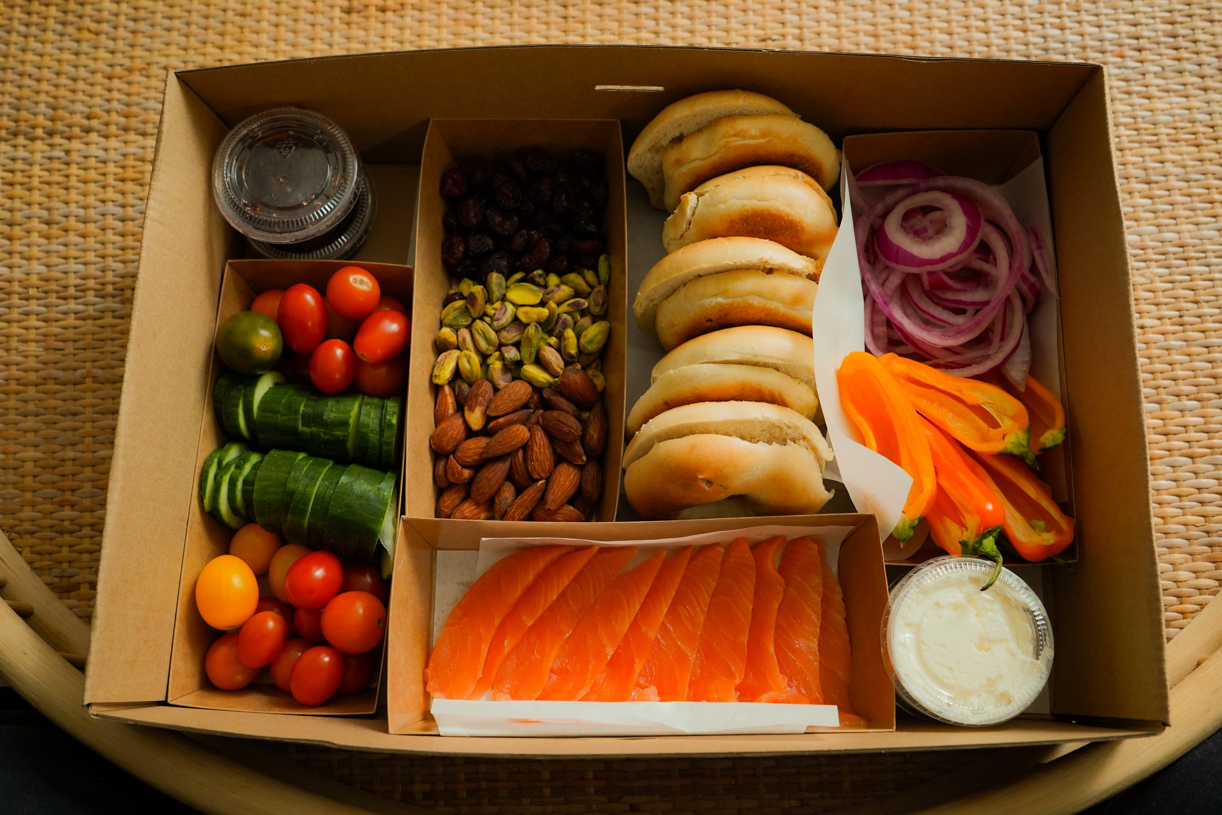 A cardboard food box contains sliced smoked salmon, red onion rings, cherry tomatoes, cucumber slices, mixed nuts, dried cranberries, pistachios, smoked turkey, and a small container of dipping sauce, on a woven placemat.