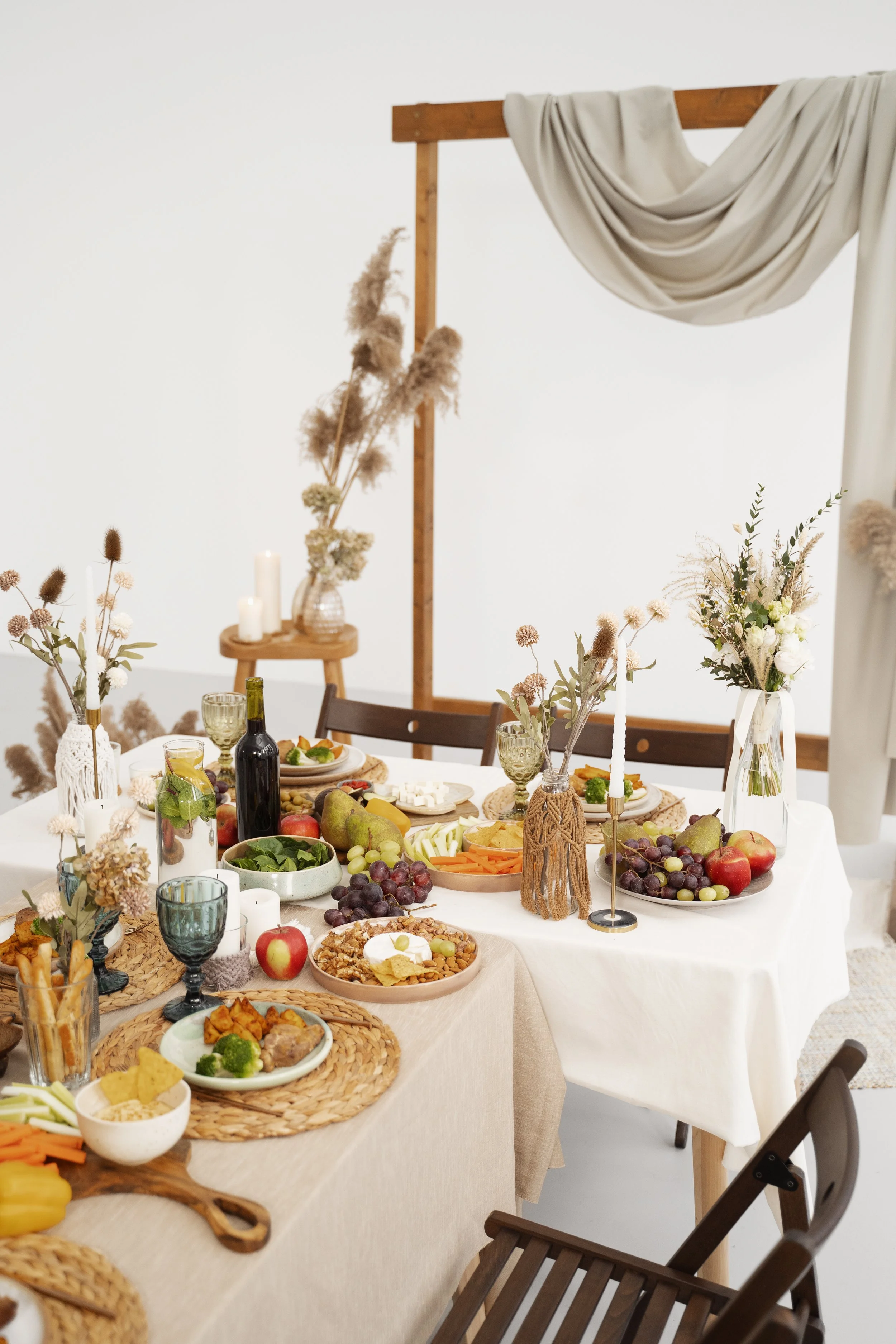 A table set for a meal with various foods including grapes, apples, vegetables, and cheese, and decorated with candles, vases with dried flowers, and woven placemats, in a bright room with a white wall and a wooden frame with draped fabric overhead.