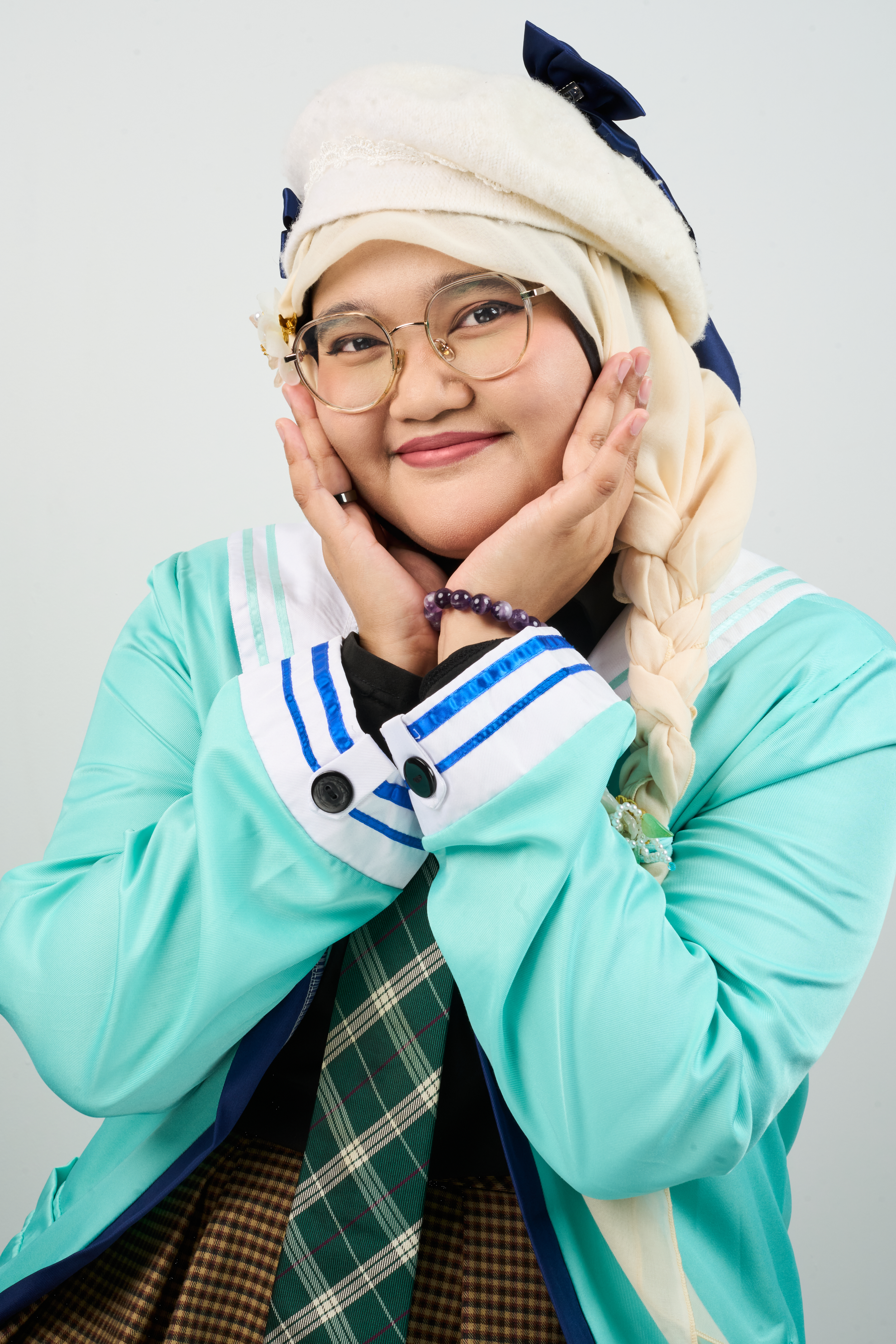 A woman wearing glasses in a teal jacket, with green tie, wearing a light yellow hijab over a white background