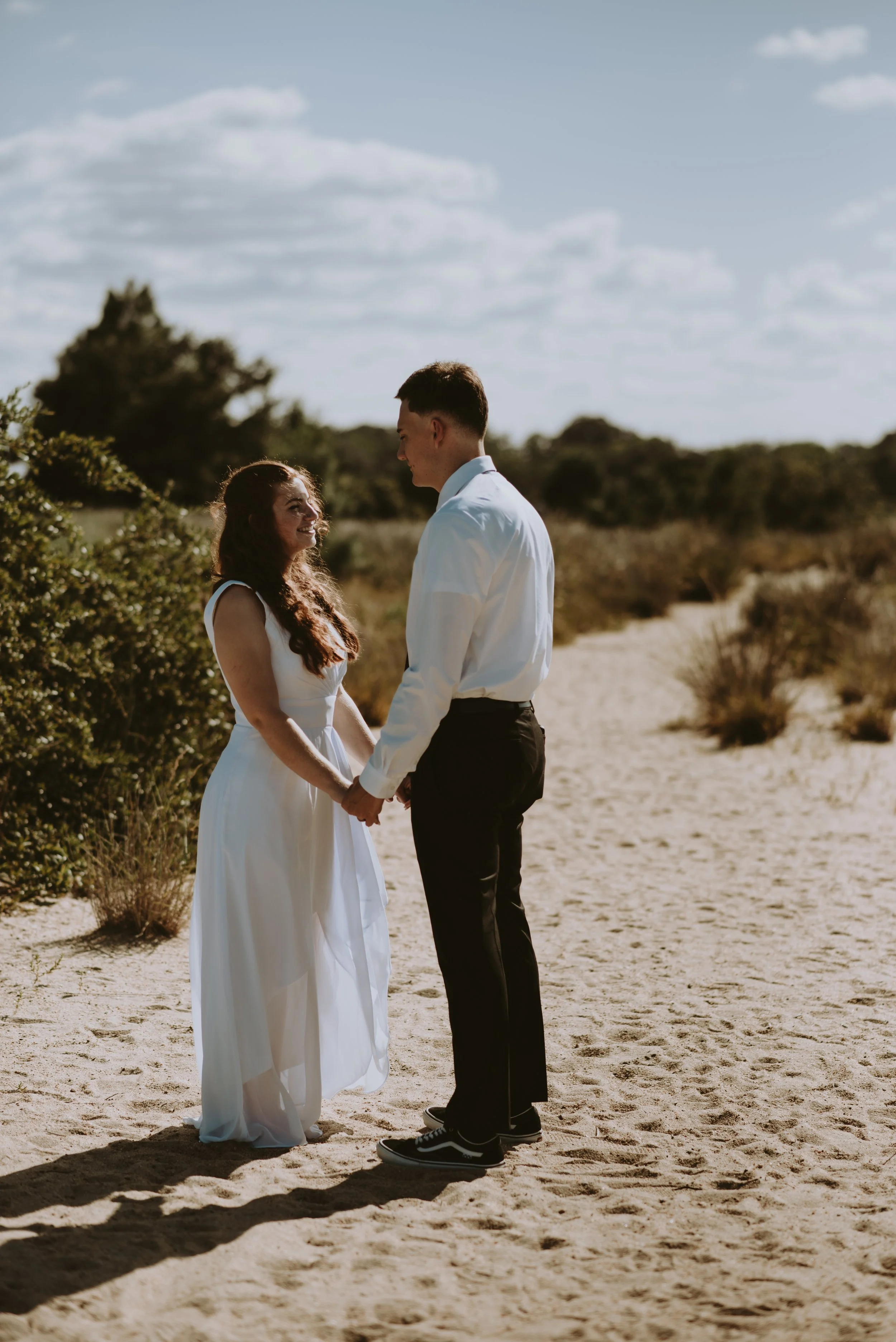 A couple dressed in wedding attire holding hands and smiling at each other on a sandy trail in a natural outdoor setting under a partly cloudy sky.