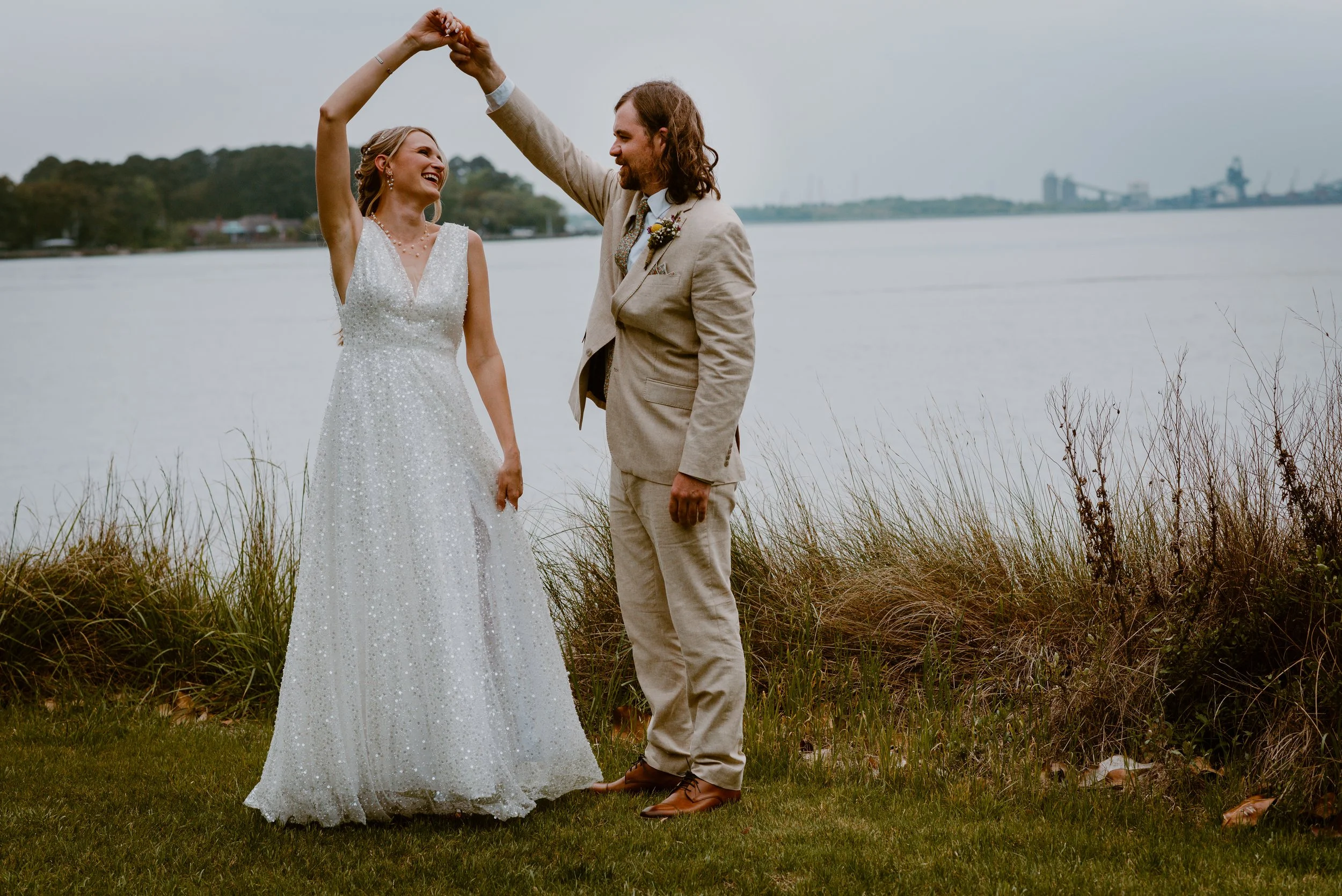 A bride and groom dancing by a body of water on their wedding day, with the groom twirling the bride while smiling.