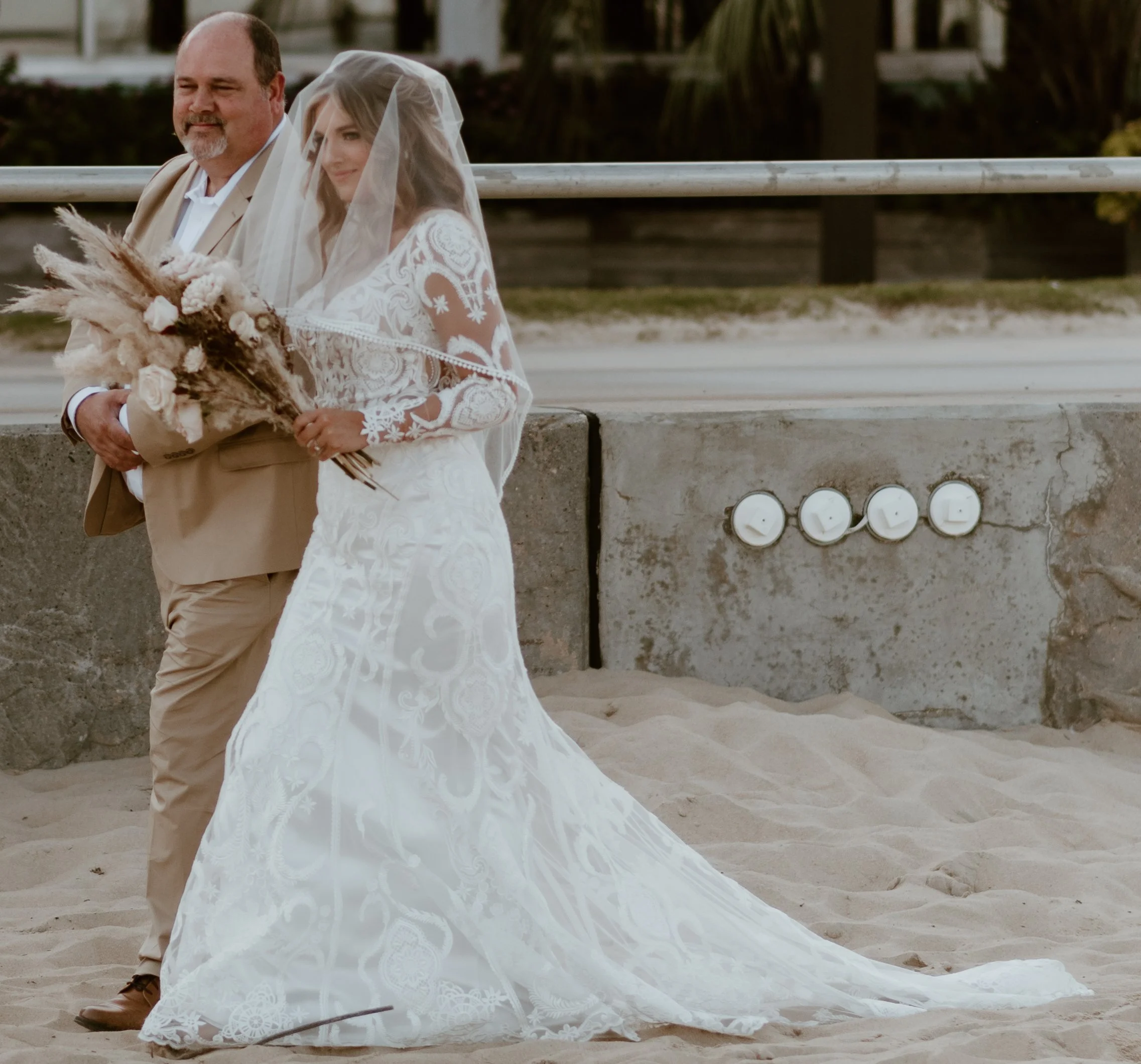 A bride in a white lace dress holding a bouquet, walking with a man in a beige suit on a sandy area near a concrete wall with four round white lights, outdoors with palm trees in the background.