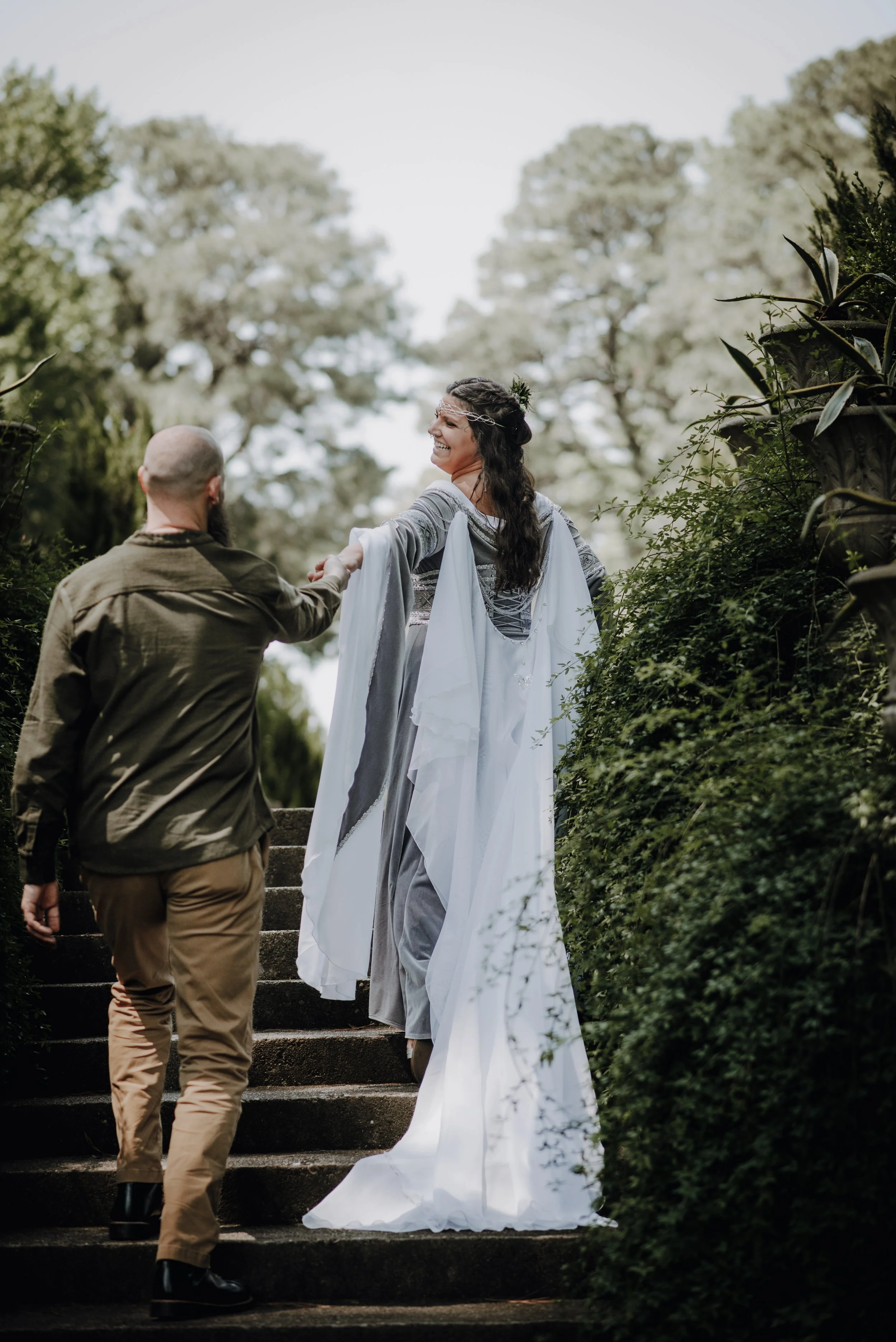 A woman in a flowing white and silver dress, smiling and reaching out to a man with a beard and shaved head, as they walk up outdoor stone steps surrounded by greenery and trees.