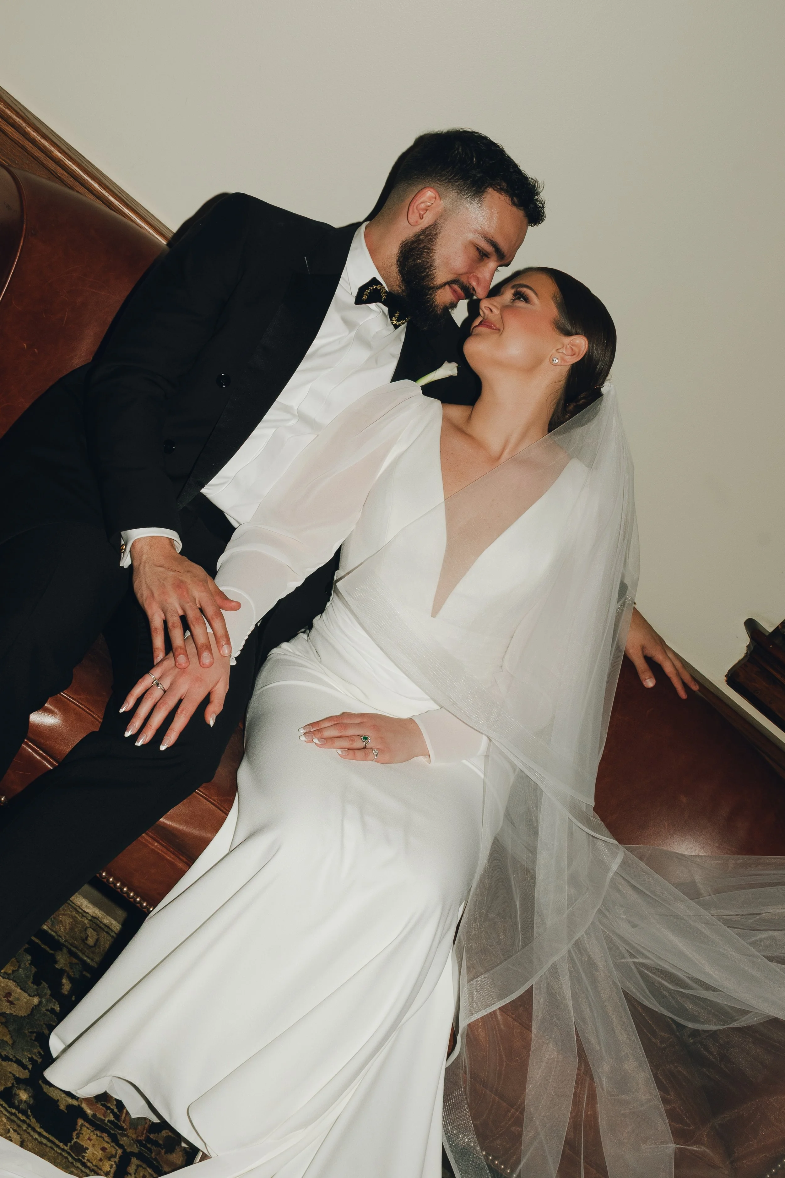 A bride and groom sitting on a brown leather sofa, leaning close with noses touching, smiling at each other during their wedding.