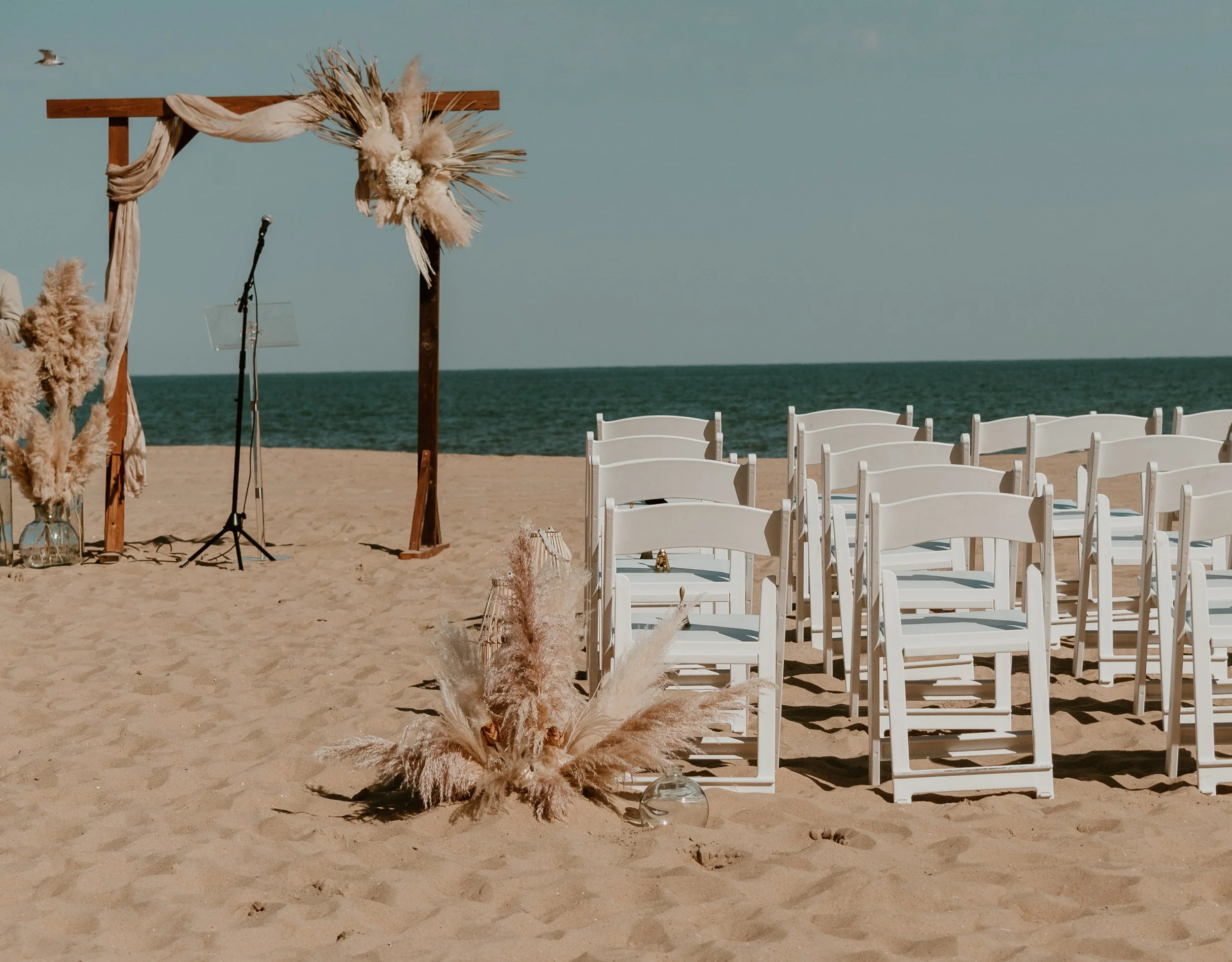 Empty white chairs set up on a sandy beach for a wedding ceremony, with a wooden arch decorated with neutral-toned fabric and pampas grass, ocean in the background, and clear blue sky.