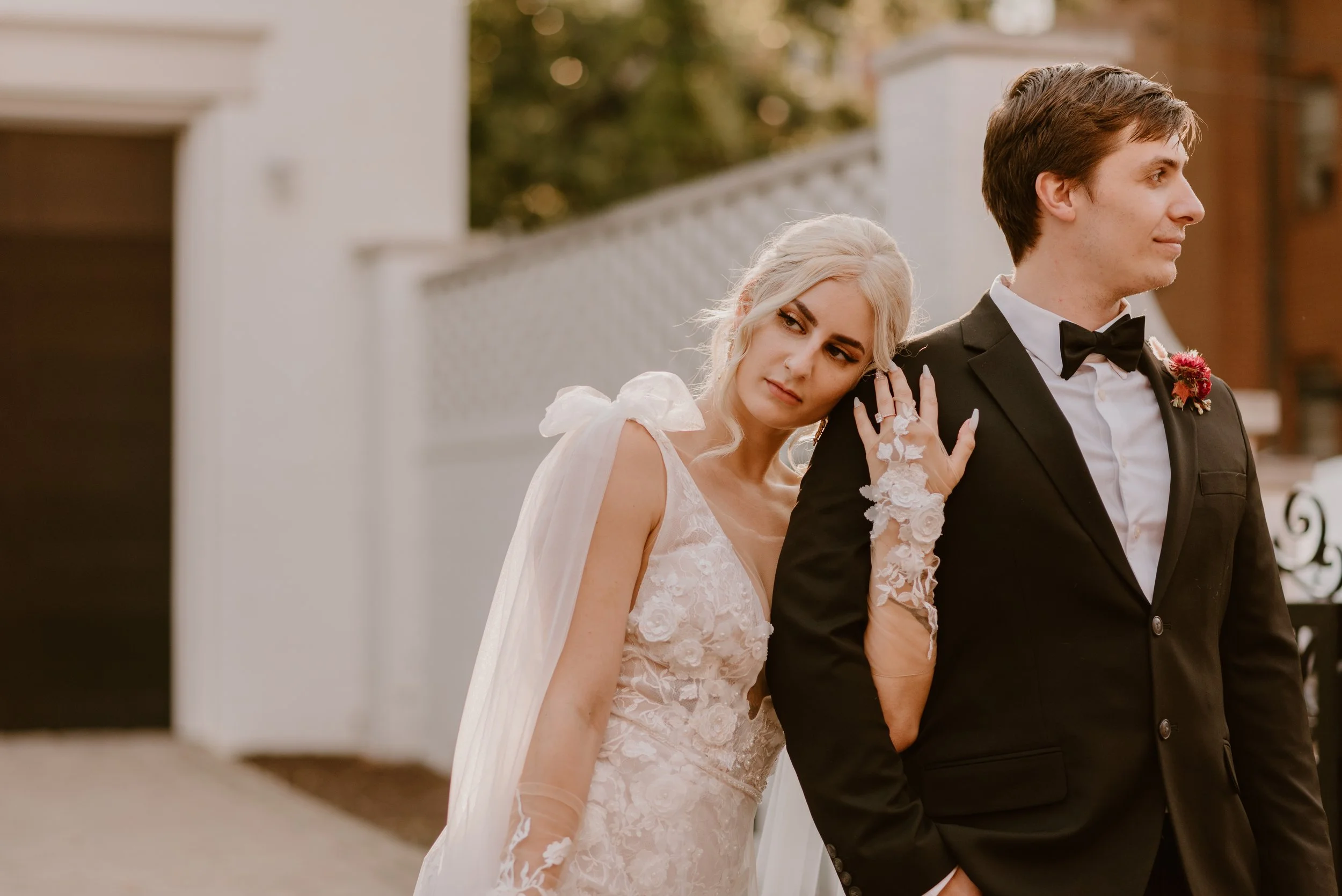 Bride leaning on groom's shoulder during wedding photos, outdoors in warm lighting.