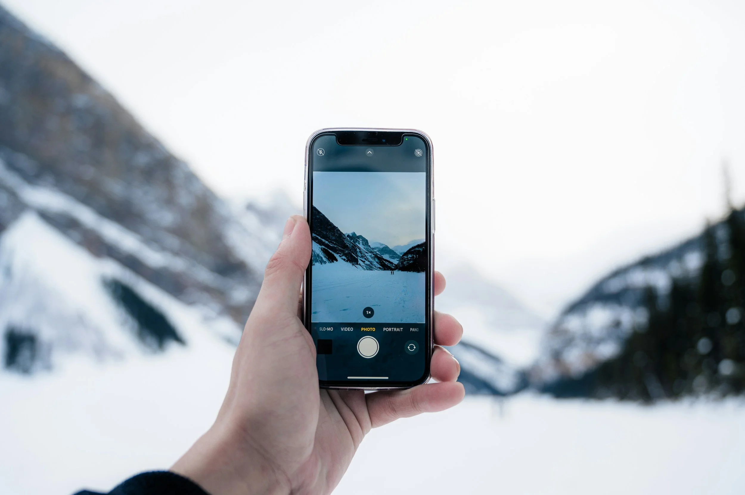 Person holding a smartphone taking a photo of a snowy mountain landscape with snow-covered mountains and a cloudy sky.