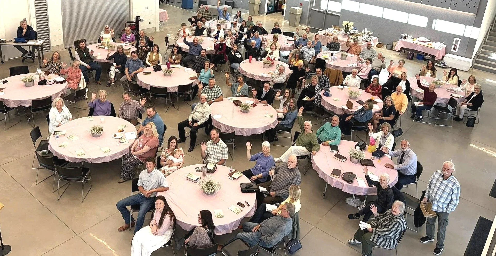 A large group of people, including men and women of various ages, sitting and standing around round tables covered with pink tablecloths inside a spacious hall, waving and smiling at the camera during a gathering or event.