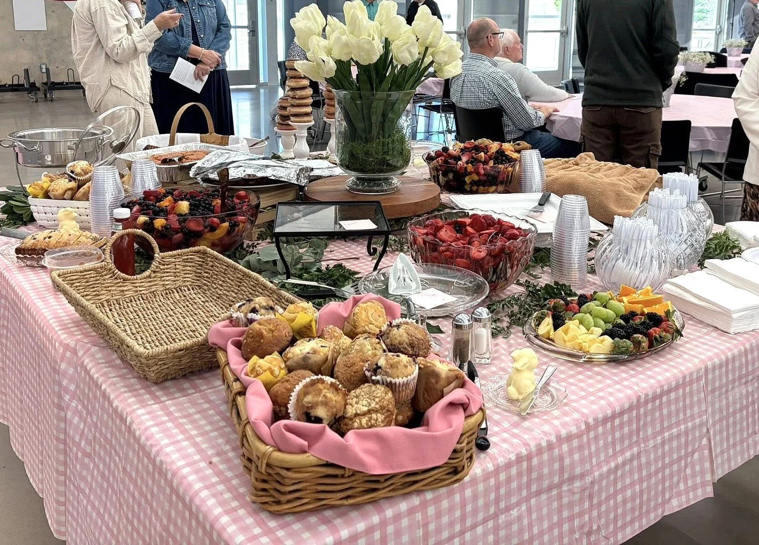 A table covered with a pink checkered tablecloth, displaying assorted baked goods, bowls of fresh berries, a vase of white tulips, and other desserts at the Lakeview Fellowship Easter Breakfast.