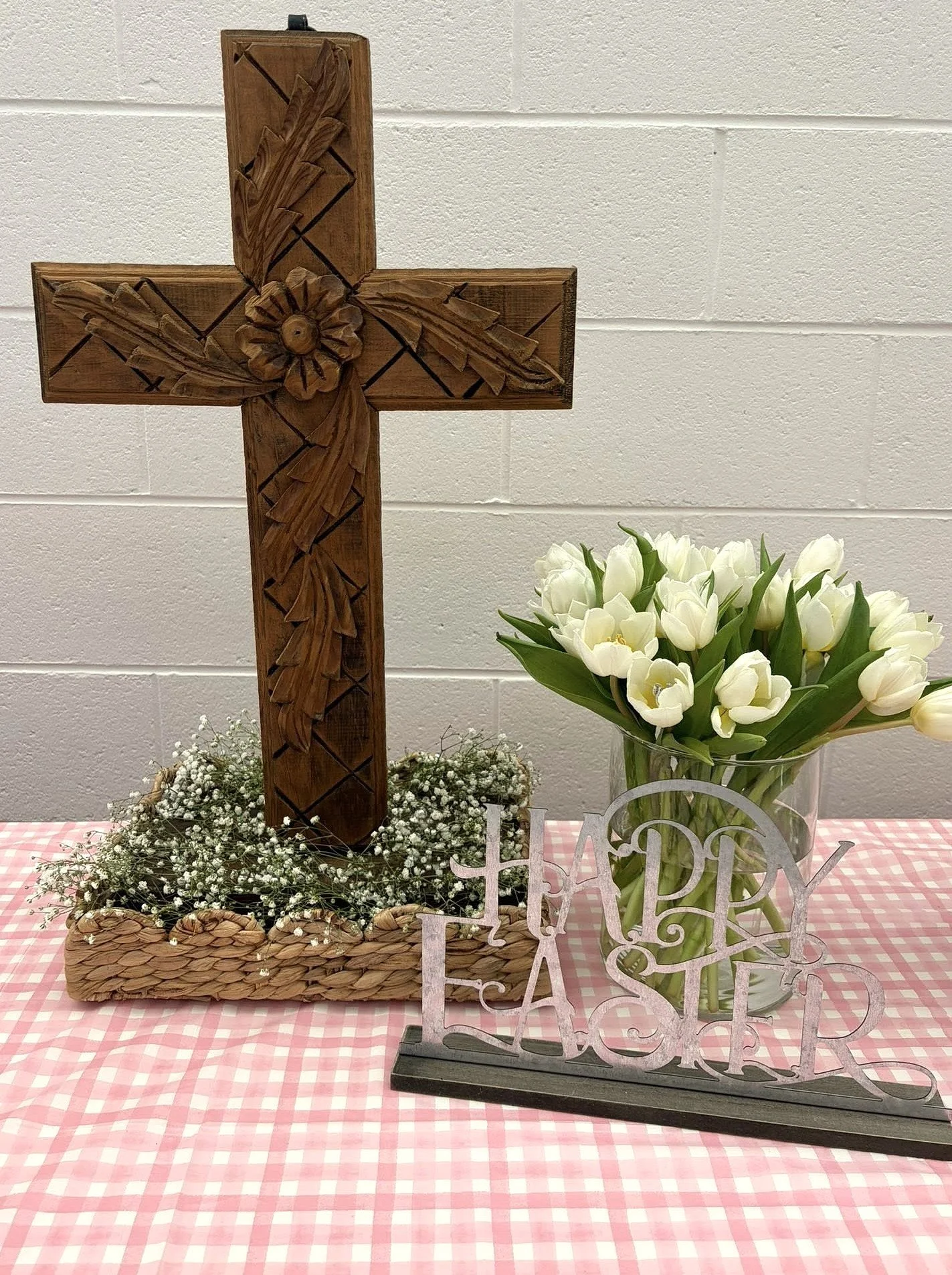 Wooden cross carved with floral design, placed on a woven basket with baby’s breath flowers, next to a glass vase filled with white tulips, and a decorative 'Happy Easter' sign on a pink checkered tablecloth.
