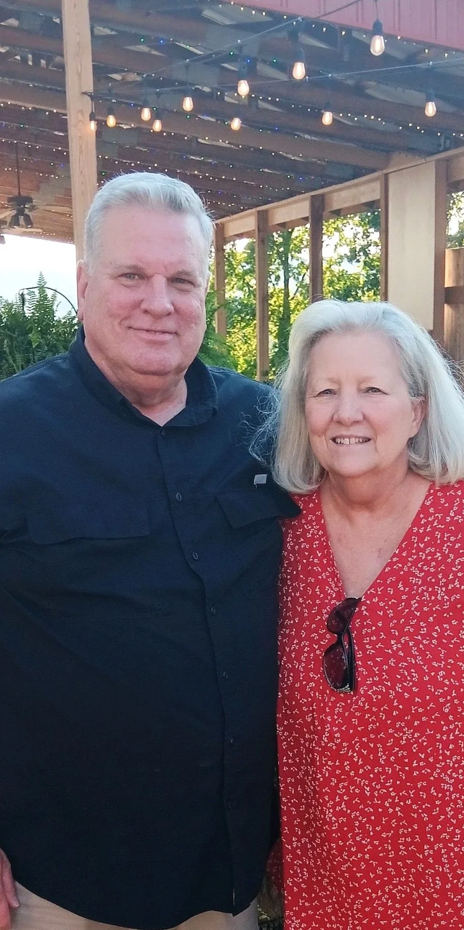 Pastor Randy and his wife smiling outdoors at the 2025 Fall Festival under string lights, with greenery in the background.